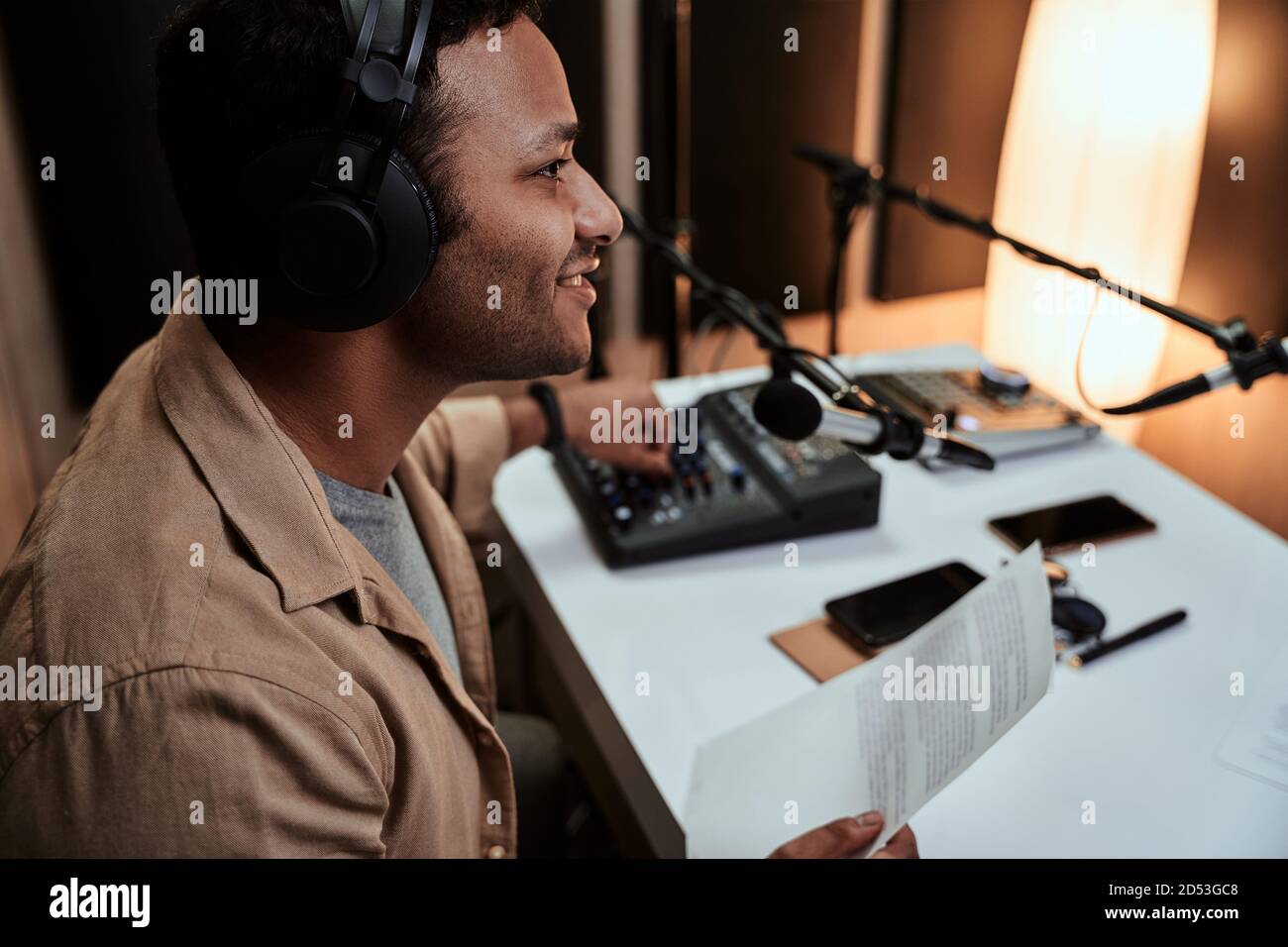Retrato de un joven presentador de radio masculino en vivo en el aire, hablando en micrófono, sosteniendo un papel de guión mientras estaba sentado en el estudio Foto de stock