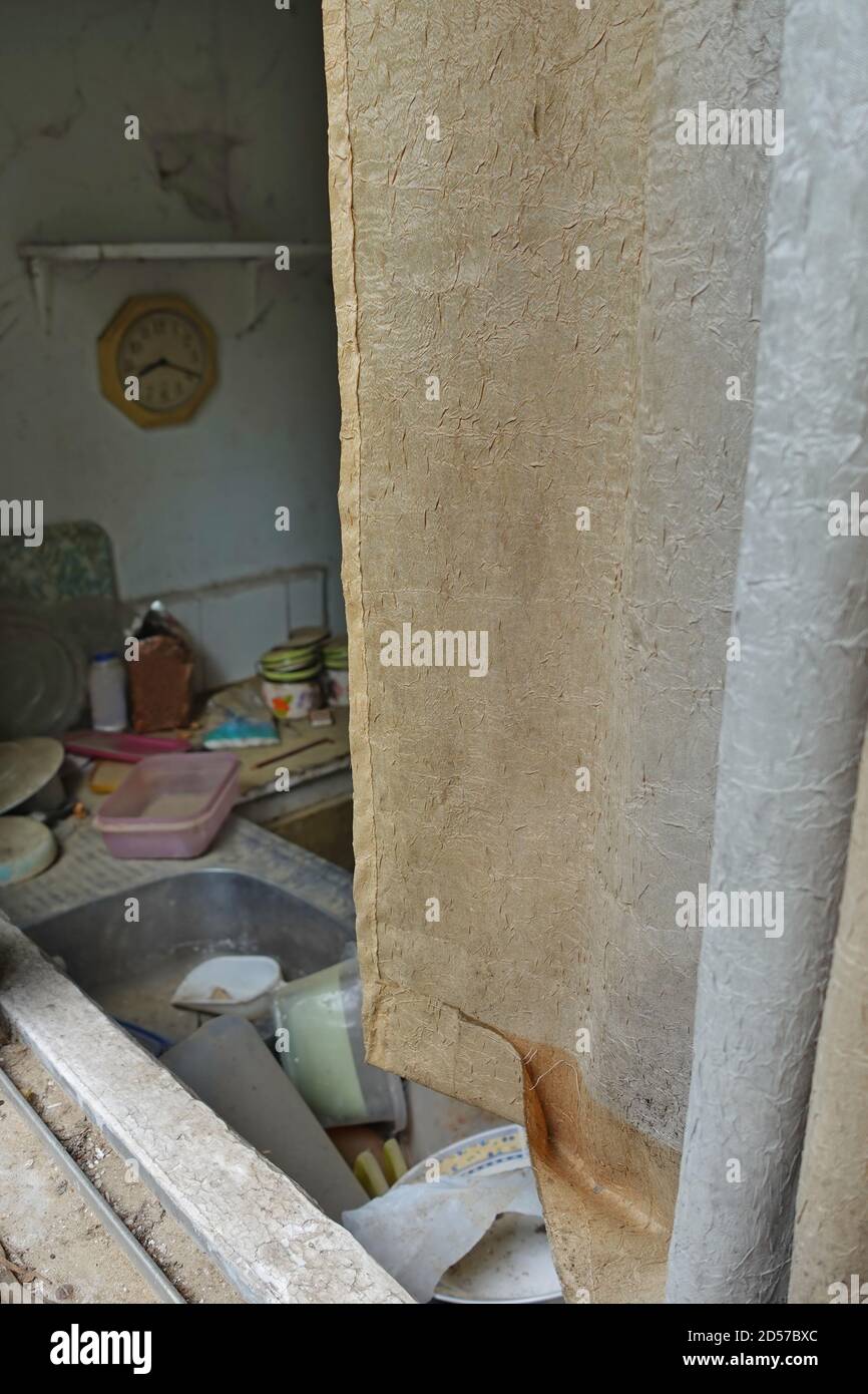 Cortina sucia y fregadero de la cocina con platos sin lavar en casa abandonada. Foto de stock
