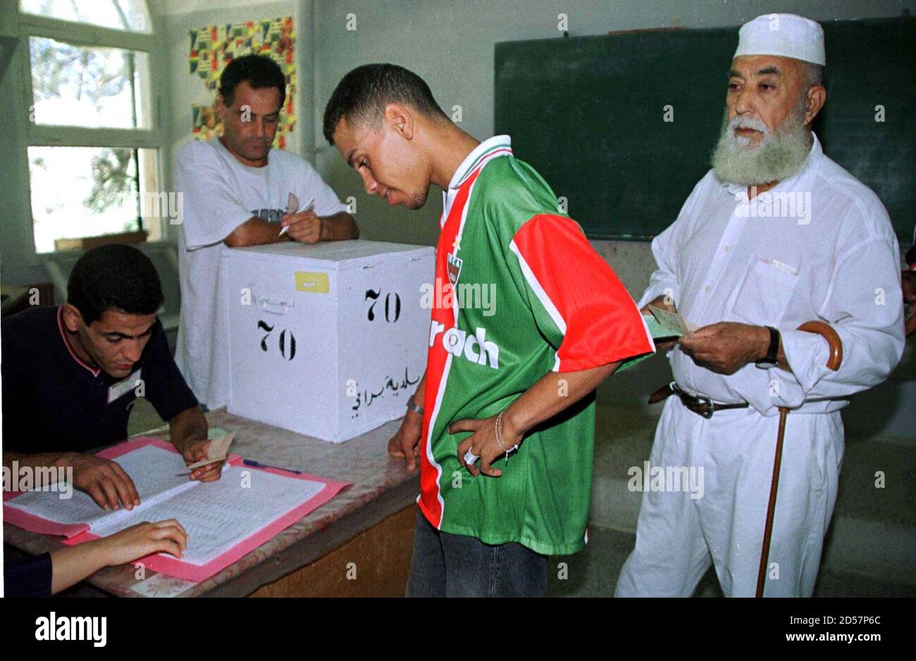Un joven argelino con los colores de un club de fútbol local se prepara para  votar como un hombre mayor con ropa tradicional espera en fila en un centro  de votación en