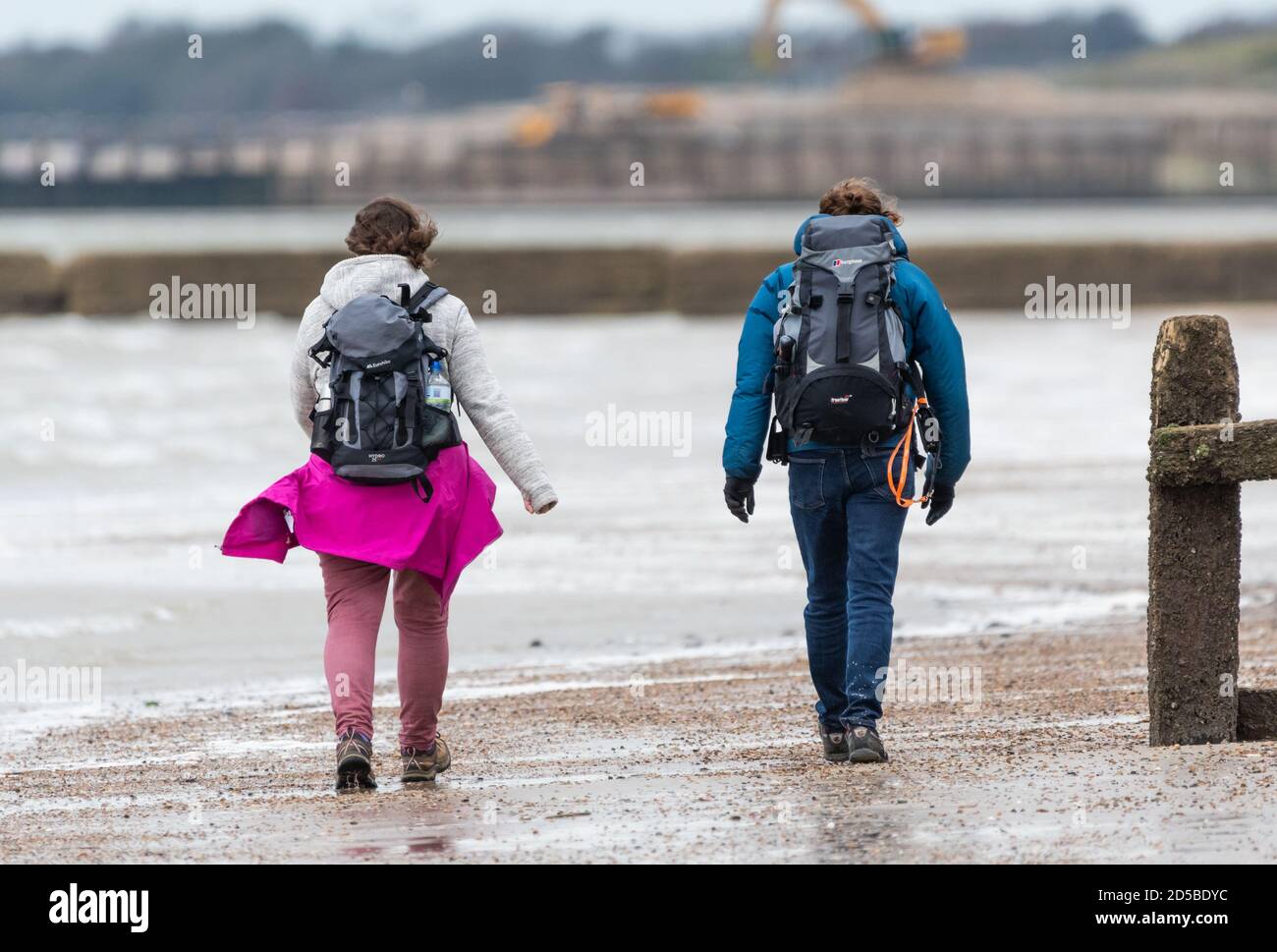 Pareja caminando en una playa con mochilas (mochilas) en clima húmedo y ventoso en el Reino Unido. Una pareja en una caminata de otoño, cada uno llevando una mochila (mochila). Foto de stock