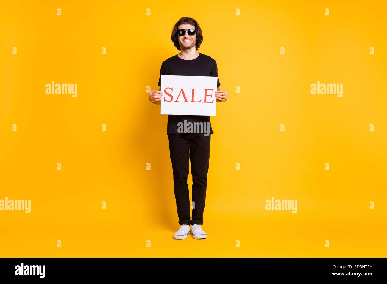 Foto de hombre joven de tamaño completo sostener la placa venta ropa  camiseta negra pantalón blanco sneakers gafas de sol aisladas amarillo  fondo de color Fotografía de stock - Alamy