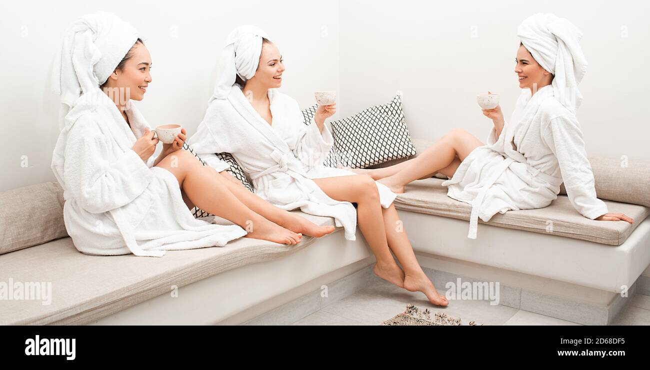 Día de té en el spa. Tres hermosas mujeres se relajan en el spa, beben té y se divierten hablando. Están vestidas con batas de baño, una toalla alrededor de sus cabezas. Foto de stock