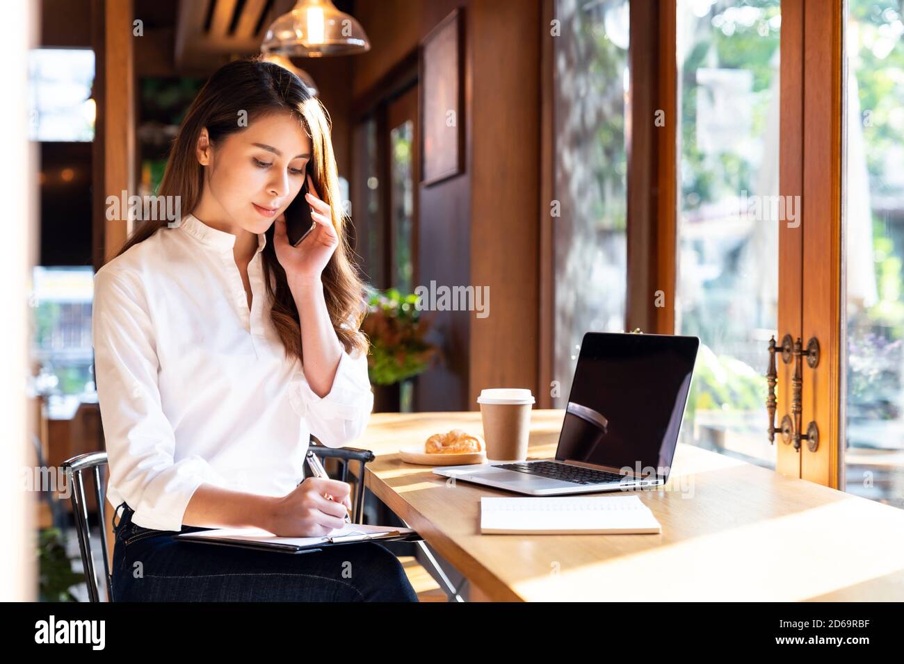Cuarentena joven mujer asiática usando ropa informal trabajo en casa en la sala de estar con ordenador portátil y beber café caliente y croissant mientras que la ciudad Fotografía de stock -
