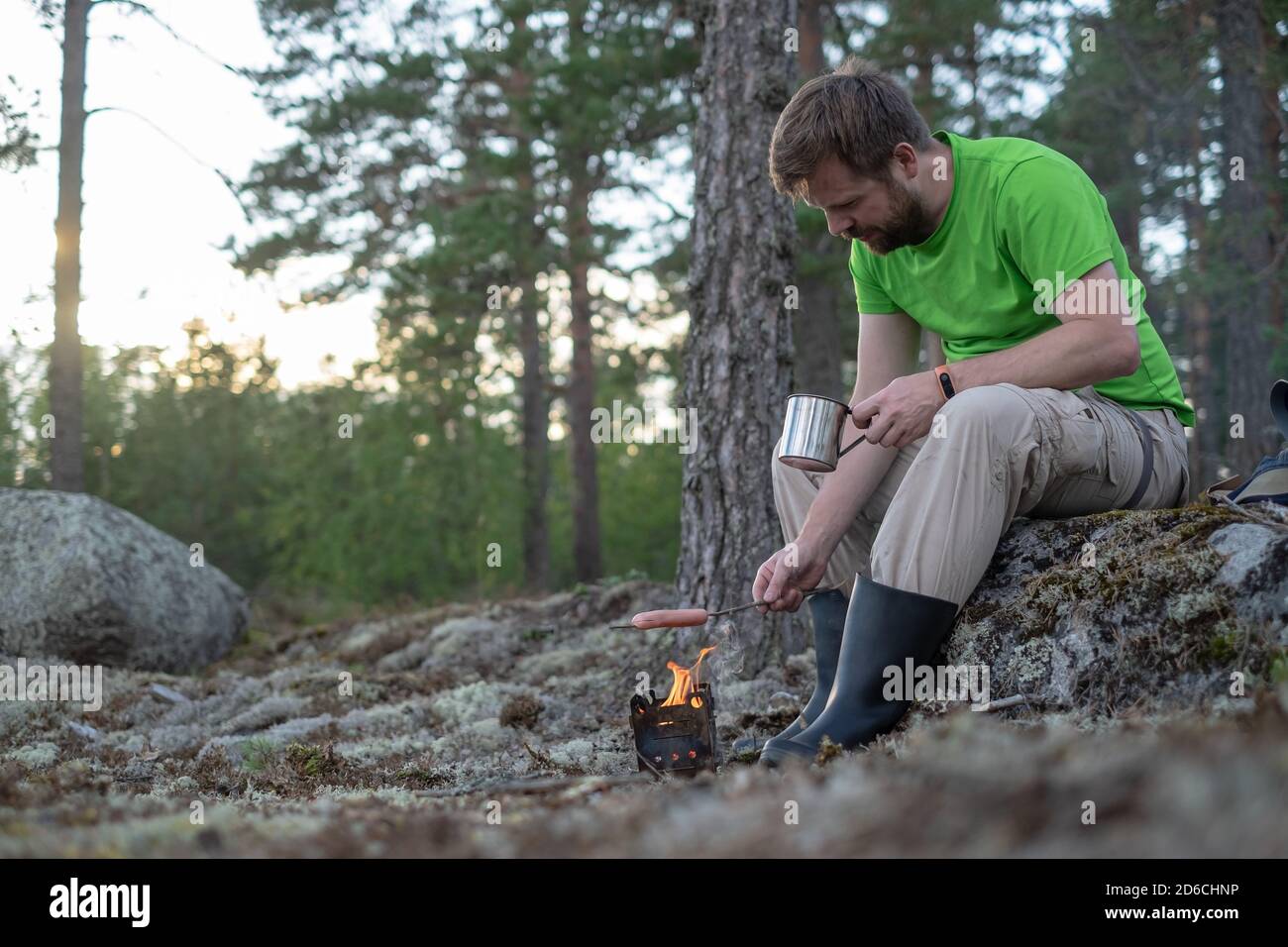El hombre retoma una salchicha sobre un fuego en una estufa de metal y sostiene una taza de bebida caliente en las manos. Cocina al aire libre. Foto de stock
