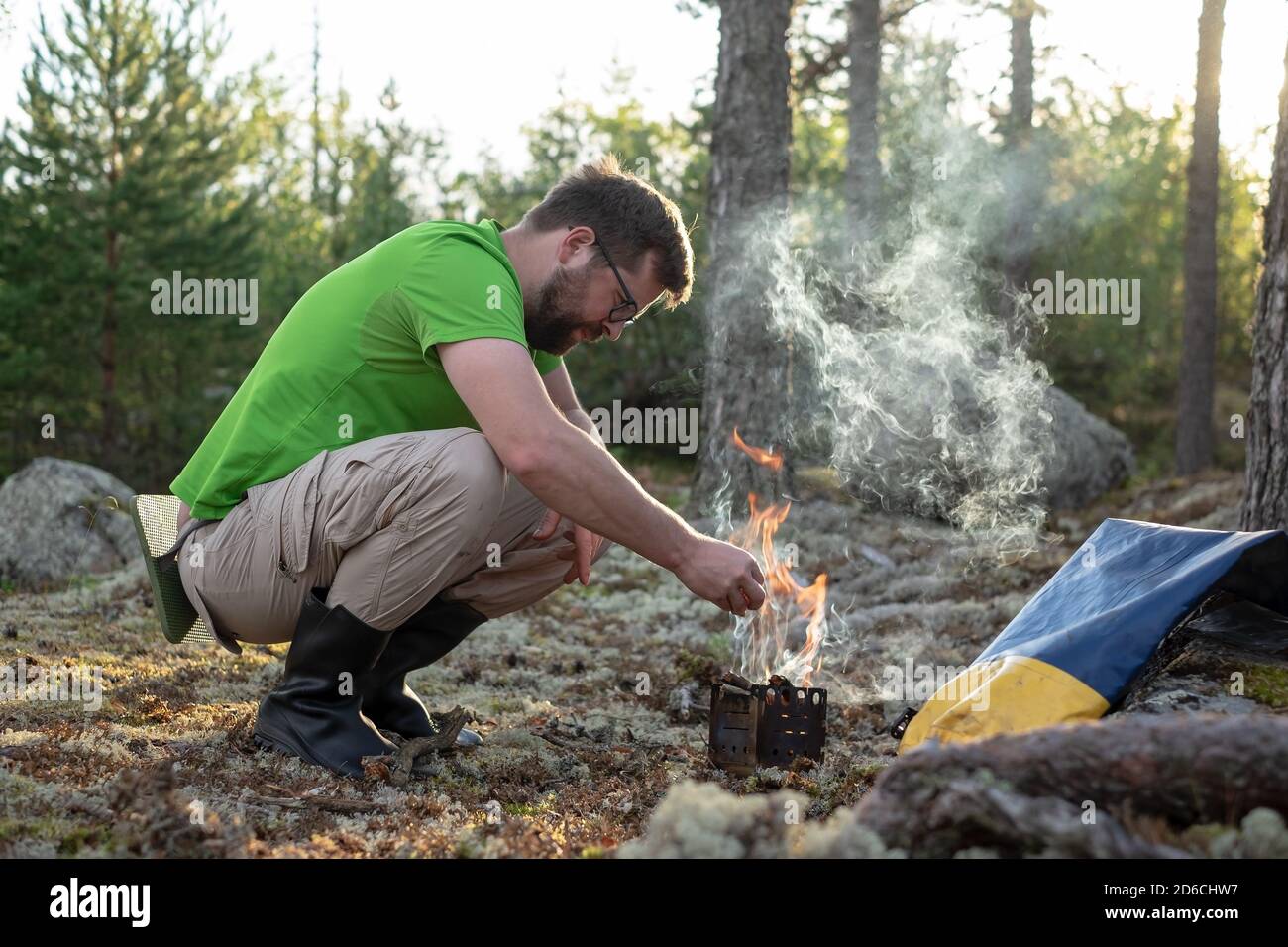 El hombre en la ropa de senderismo calienta un quemador de metal prefabricado con madera, para cocinar, en el bosque, en un campamento. Descanso activo. Foto de stock