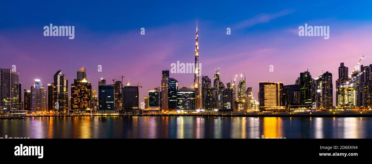 Impresionantes vistas del horizonte iluminado de Dubai al atardecer con modernos rascacielos reflejados en el canal de agua que fluye en primer plano. Foto de stock