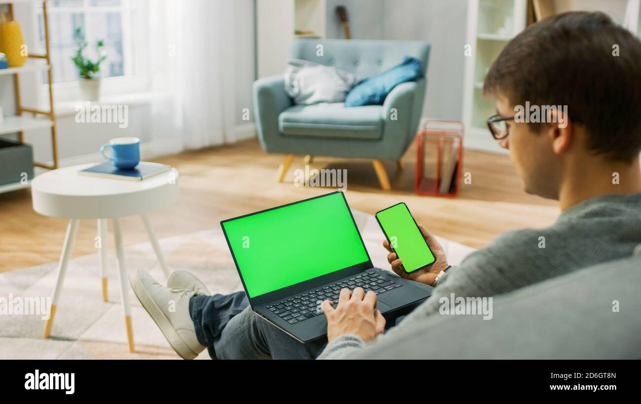 Young Man at Home trabaja en un ordenador portátil con pantalla de bloqueo verde, mientras sostiene un smartphone con pantalla Chroma Key. Está sentado en un sofá Foto de stock