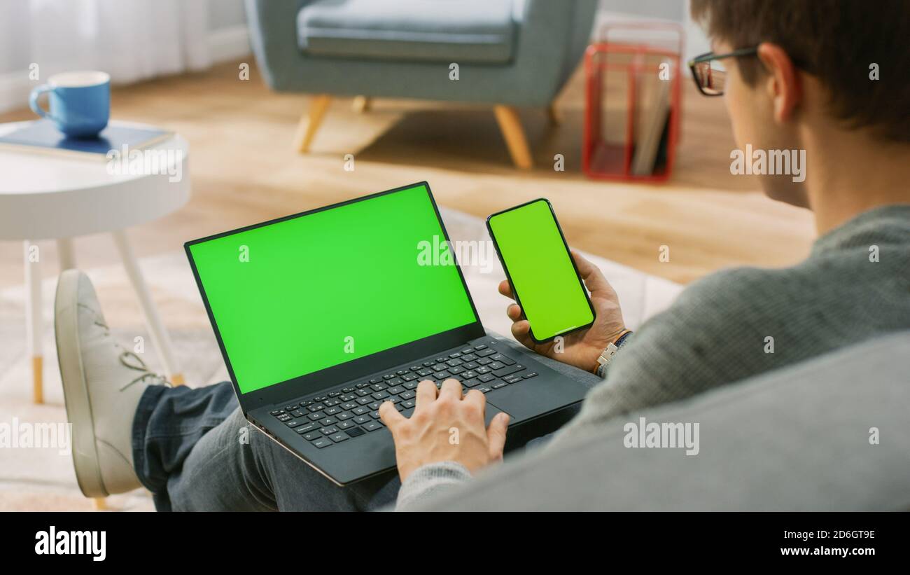 Young Man at Home trabaja en un ordenador portátil con pantalla de bloqueo verde, mientras sostiene un smartphone con pantalla Chroma Key. Está sentado en un sofá Foto de stock