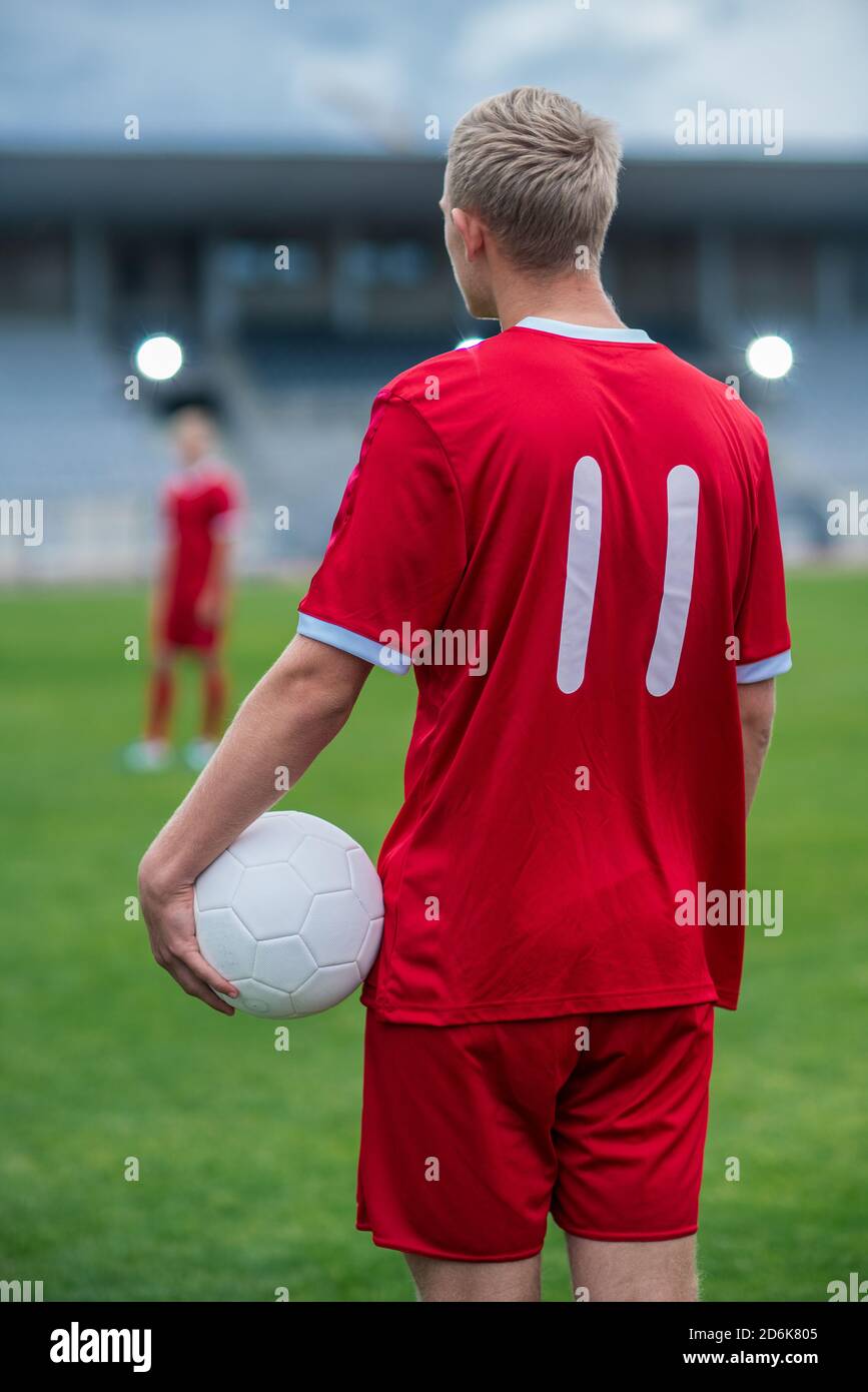 Back Shot de un jugador de fútbol profesional que se encuentra en un estadio durante el partido de campeonato y que celebra una pelota de fútbol blanca. Su compañero de equipo está adentro Foto de stock