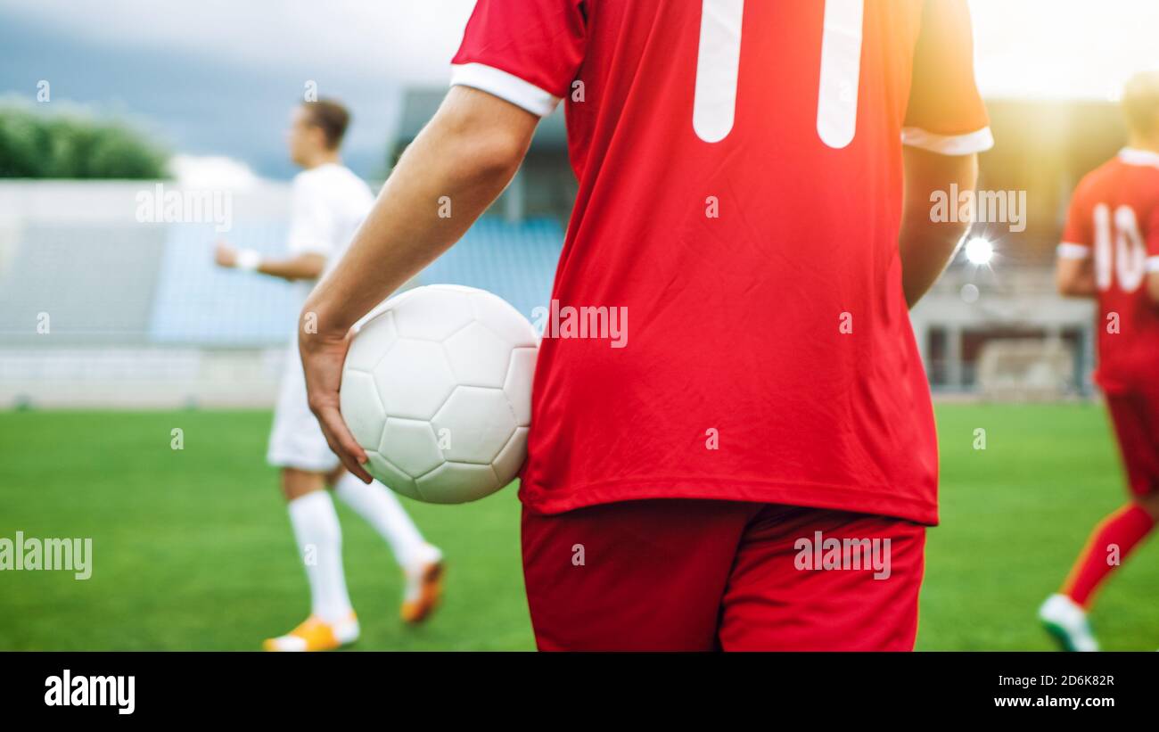 Tiro de un jugador de fútbol que lleva el balón mientras camina por el campo. En el fondo formación de equipo profesional. Foto de stock