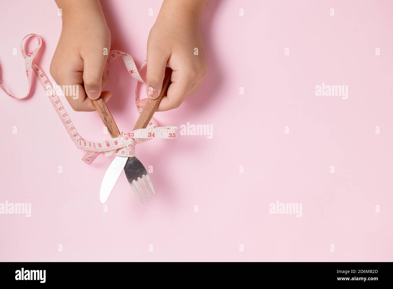 Gordo niño mano sosteniendo cuchillo y tenedor envuelto en cinta métrica sobre fondo rosa. Concepto de dieta Foto de stock