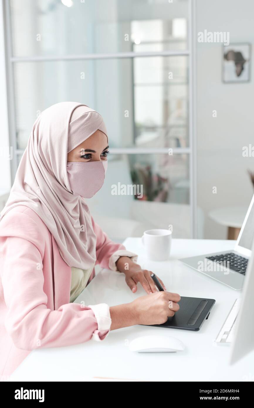 Joven diseñadora creativa en hijab y máscara protectora sentada por ordenador Foto de stock