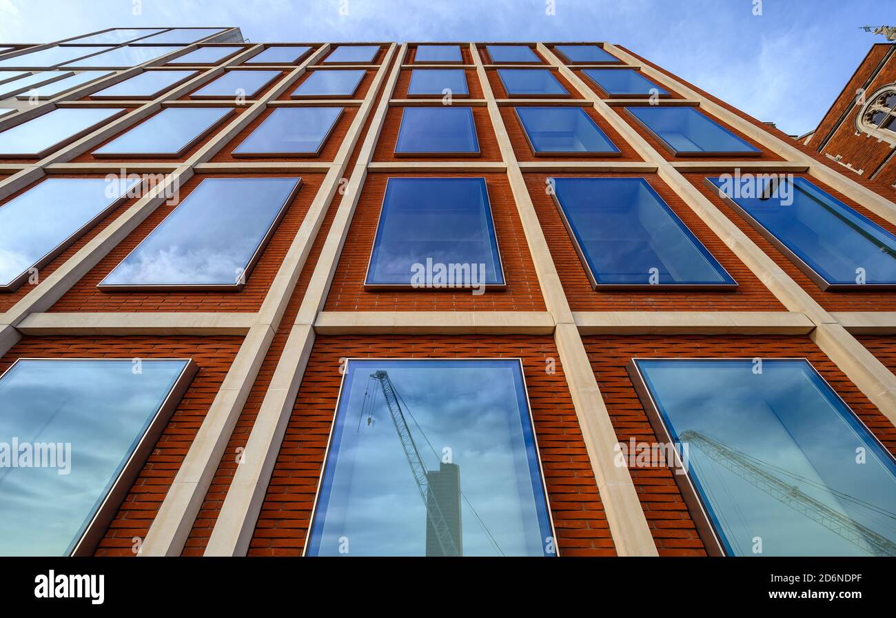 Un edificio de oficinas en Londres con ladrillos rojos y ventanas azules. Las líneas rectas dan una fuerte perspectiva hacia arriba. Reflejos de grúas en las ventanas. Foto de stock
