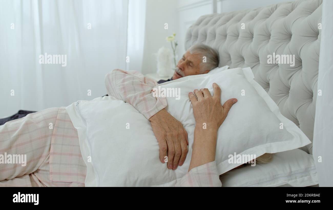 Los abuelos caucásicos mayores se unen acostados y durmiendo en la cama en  casa. Mujer irritada que se molesta con el hombre ronquidos fuerte. Abuela  cubriendo sus orejas con almohada mientras el