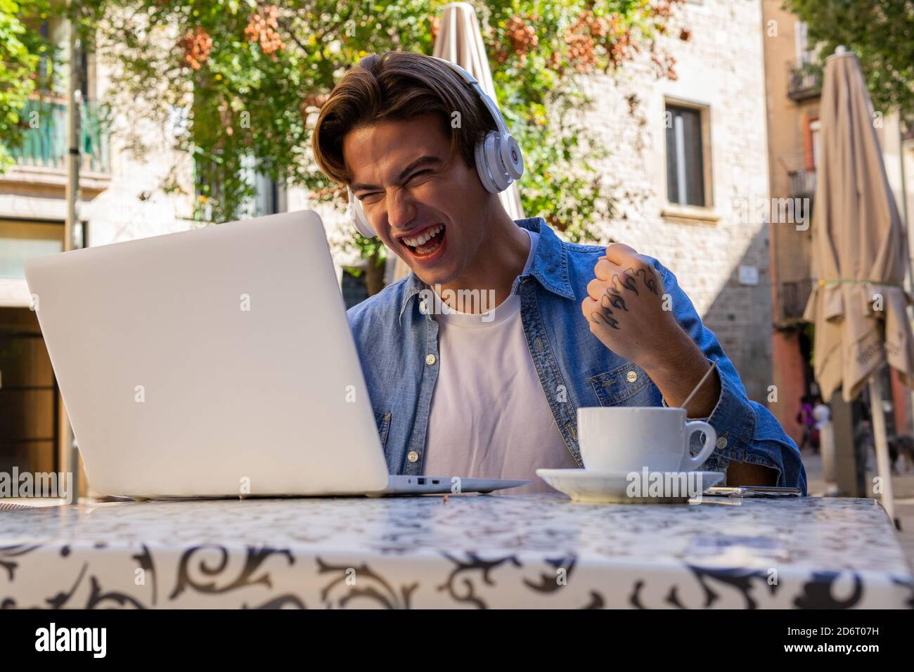Un joven demasiado alegre con unos auriculares inalámbricos recibiendo  noticias increíbles portátil y celebrar la victoria mientras se sienta en  la mesa con taza de café en el café Fotografía de stock -