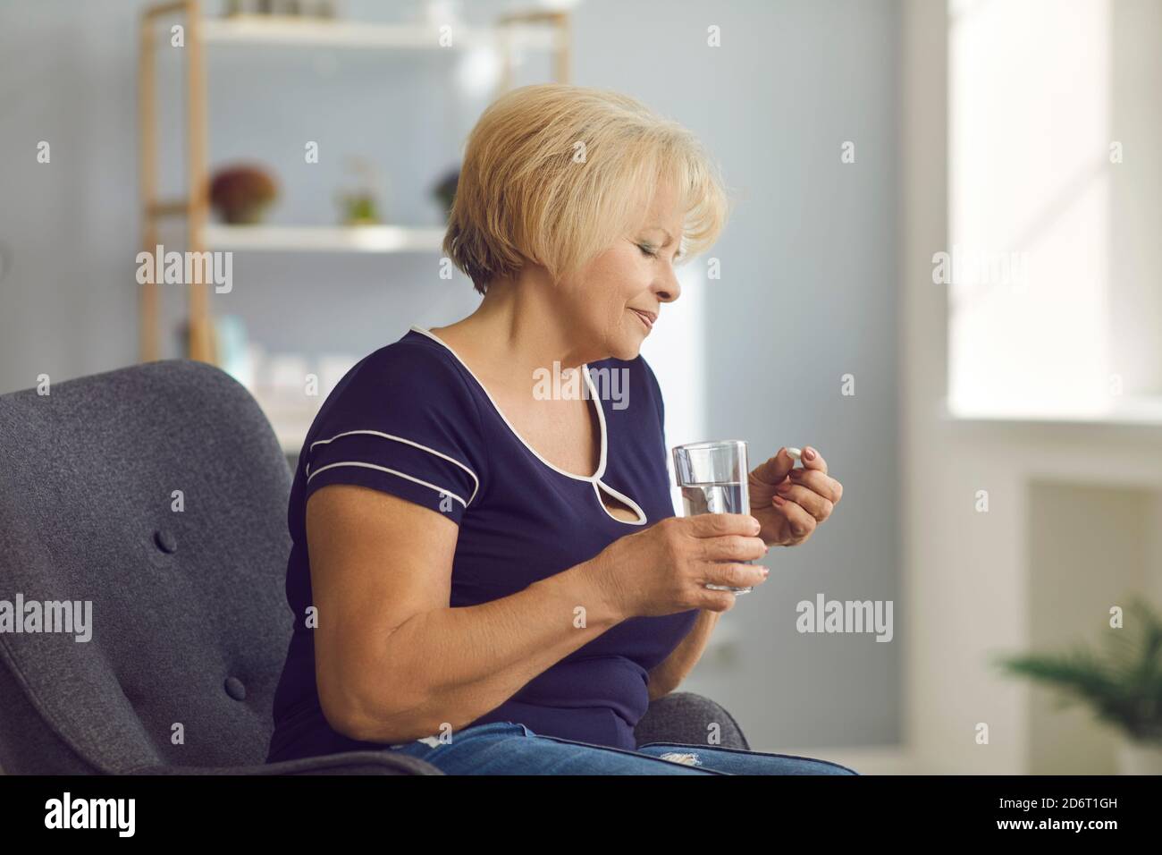 Mujer sonriente madura sentada en el sillón y tomando la píldora médica y agua pura en el cristal en las manos Foto de stock