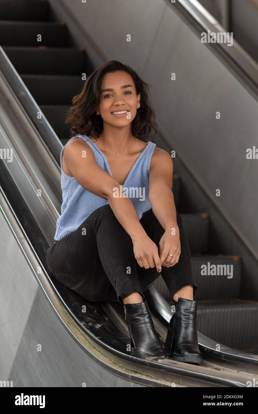 hermosa mujer latina joven con pelo ondulado corto, sentada en el pasamanos  de algunas escaleras eléctricas, lleva una blusa azul, pantalones negros y  botas, casual Fotografía de stock - Alamy