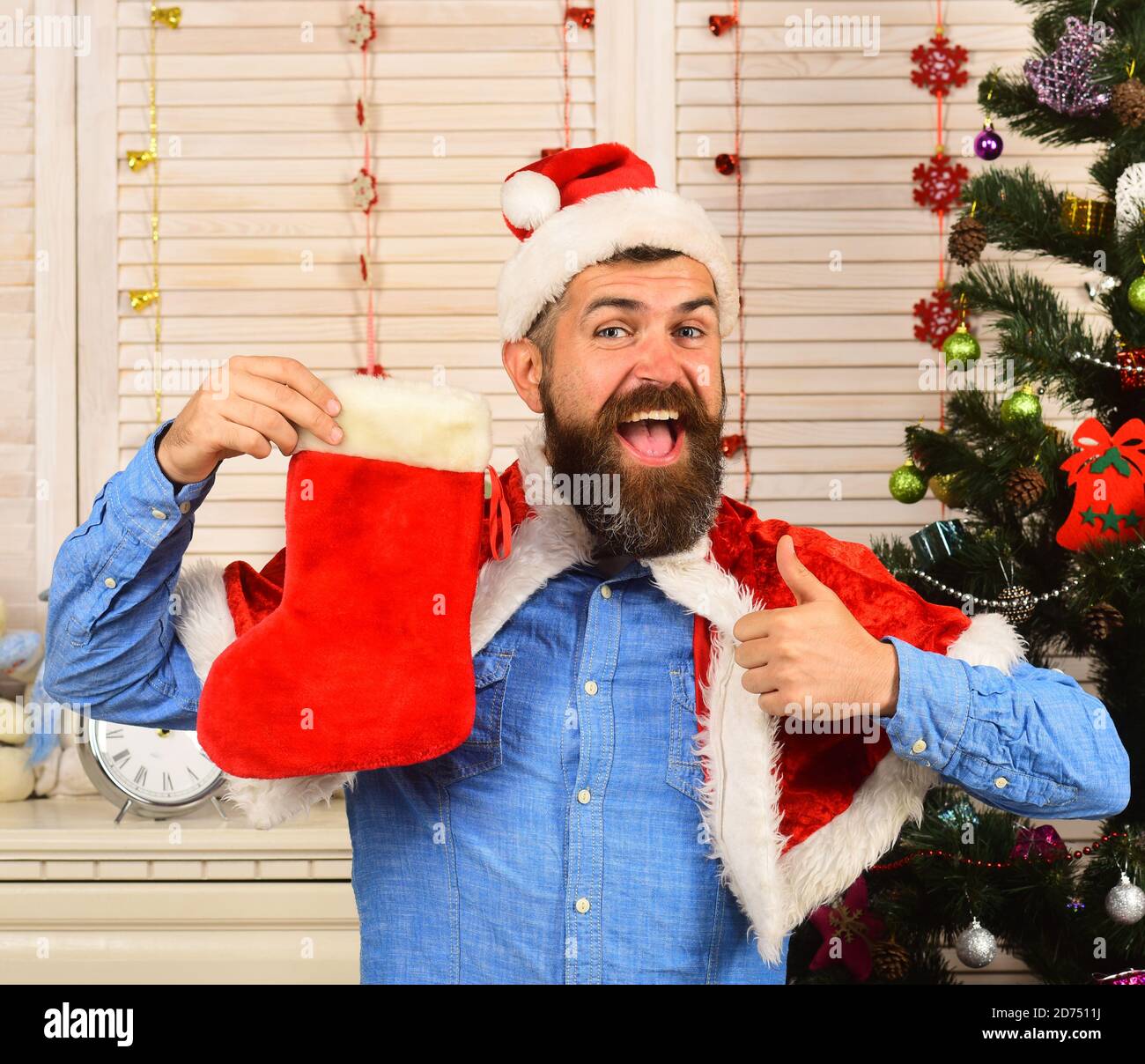 Papá Noel con cara feliz cerca del árbol de Navidad sobre fondo de madera.  Festivales y presenta el concepto. Hombre con barba en camisa azul muestra  los pulgares hacia arriba. El chico