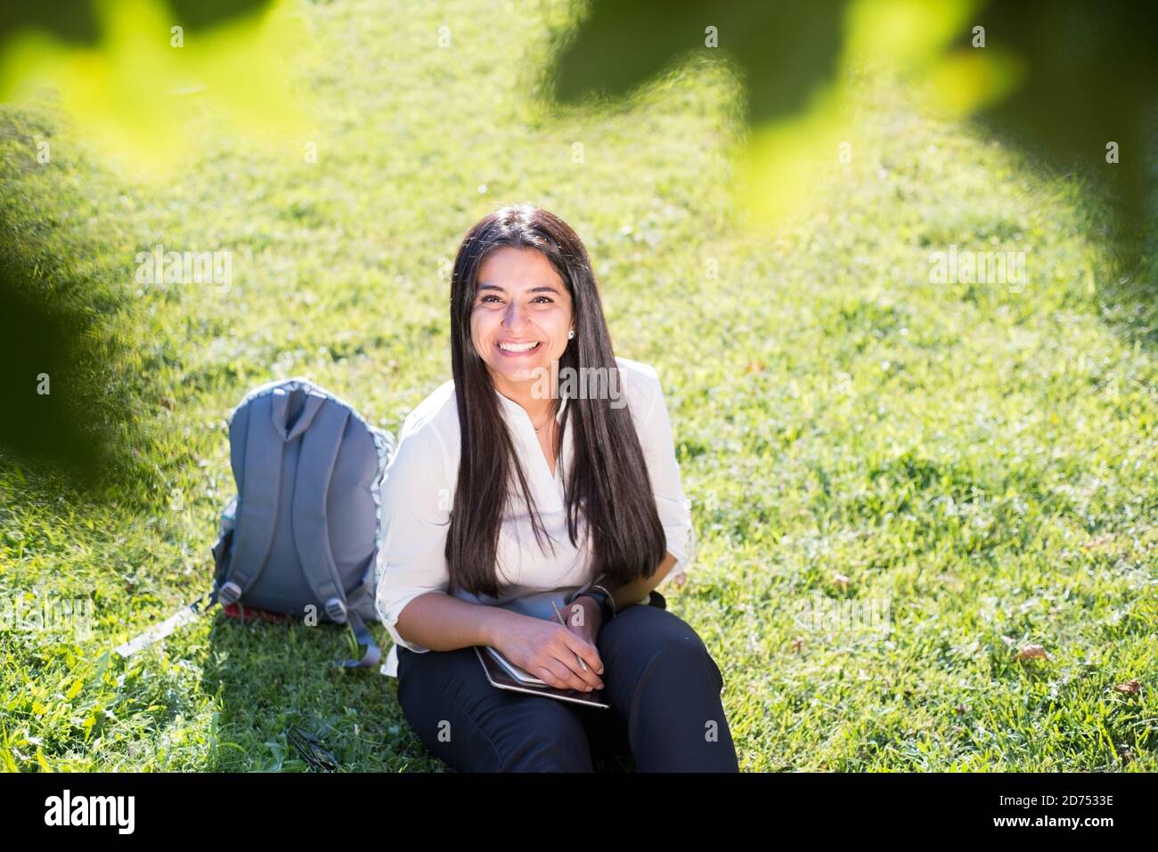 La niña India, estudiante, sonríe, se sienta en la hierba verde, con una mochila y un bloc de notas, escribe Foto de stock