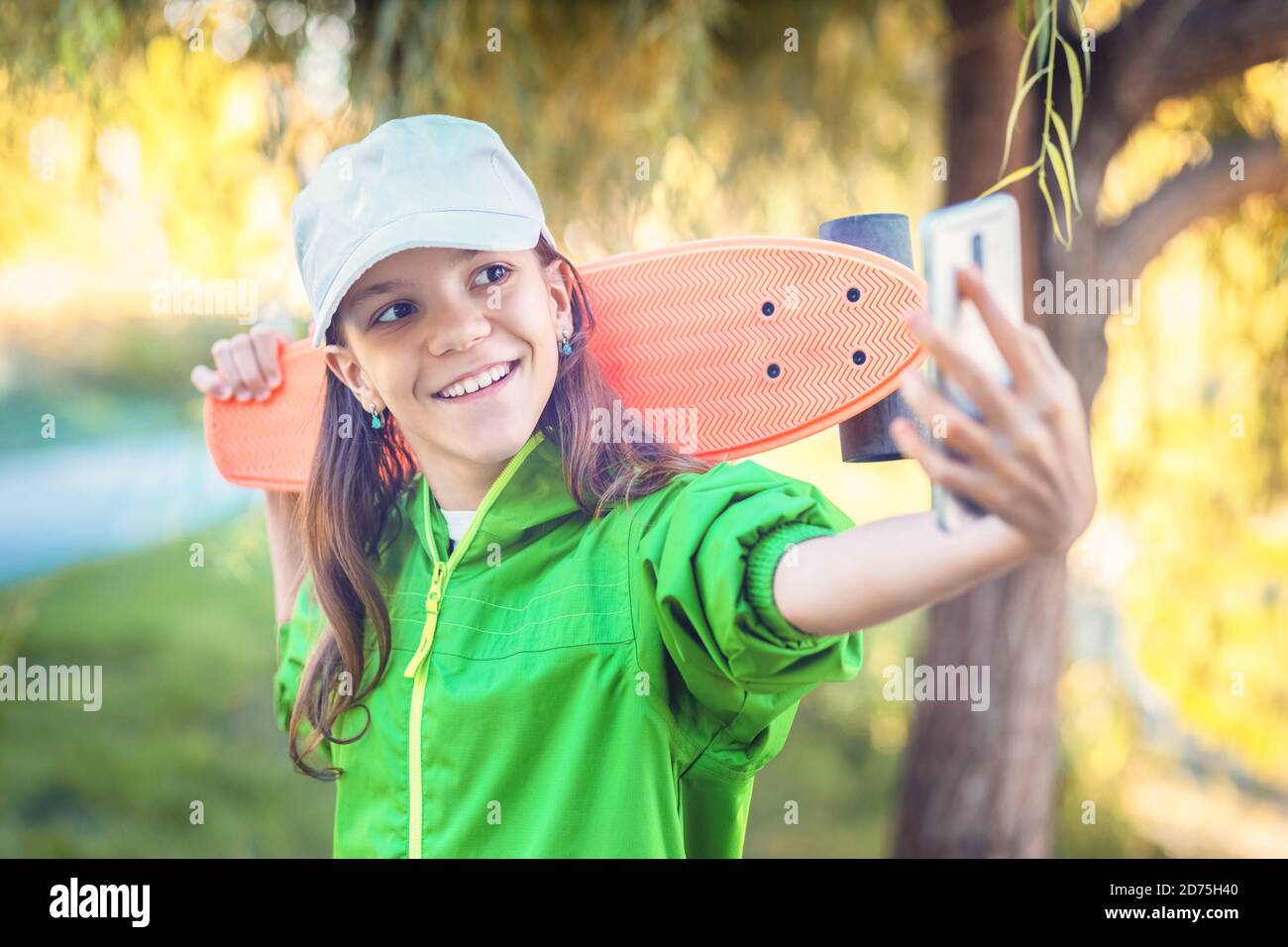Feliz niña hipster tomando selfie con el teléfono celular mientras sosteniendo monopatín Foto de stock