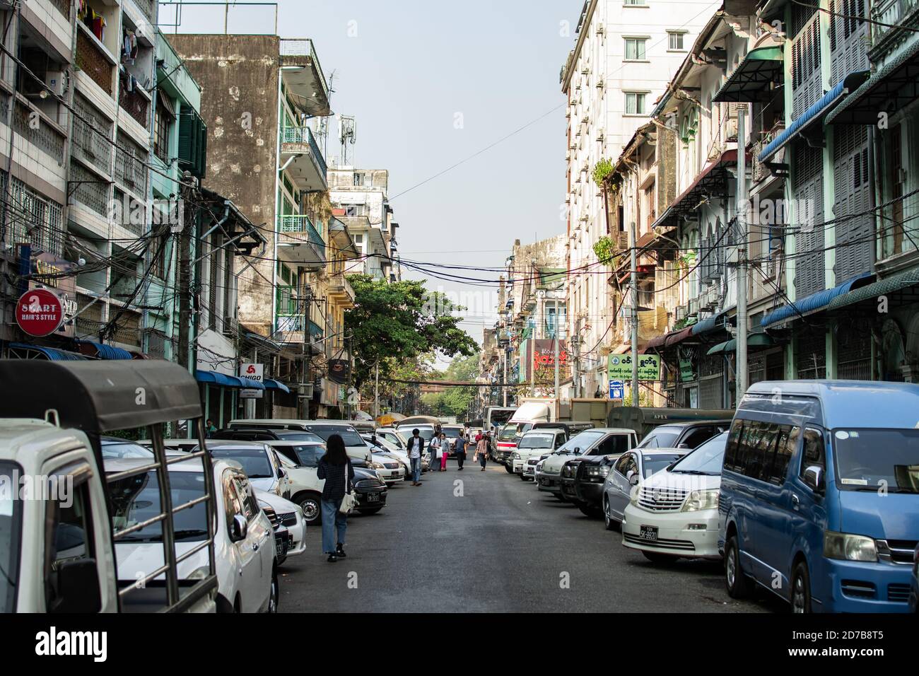Yangon, Myanmar - 30 de diciembre de 2019: La vida cotidiana ocurre en una calle estrecha local en el centro de la ciudad con coches estacionados y peatones caminando Foto de stock