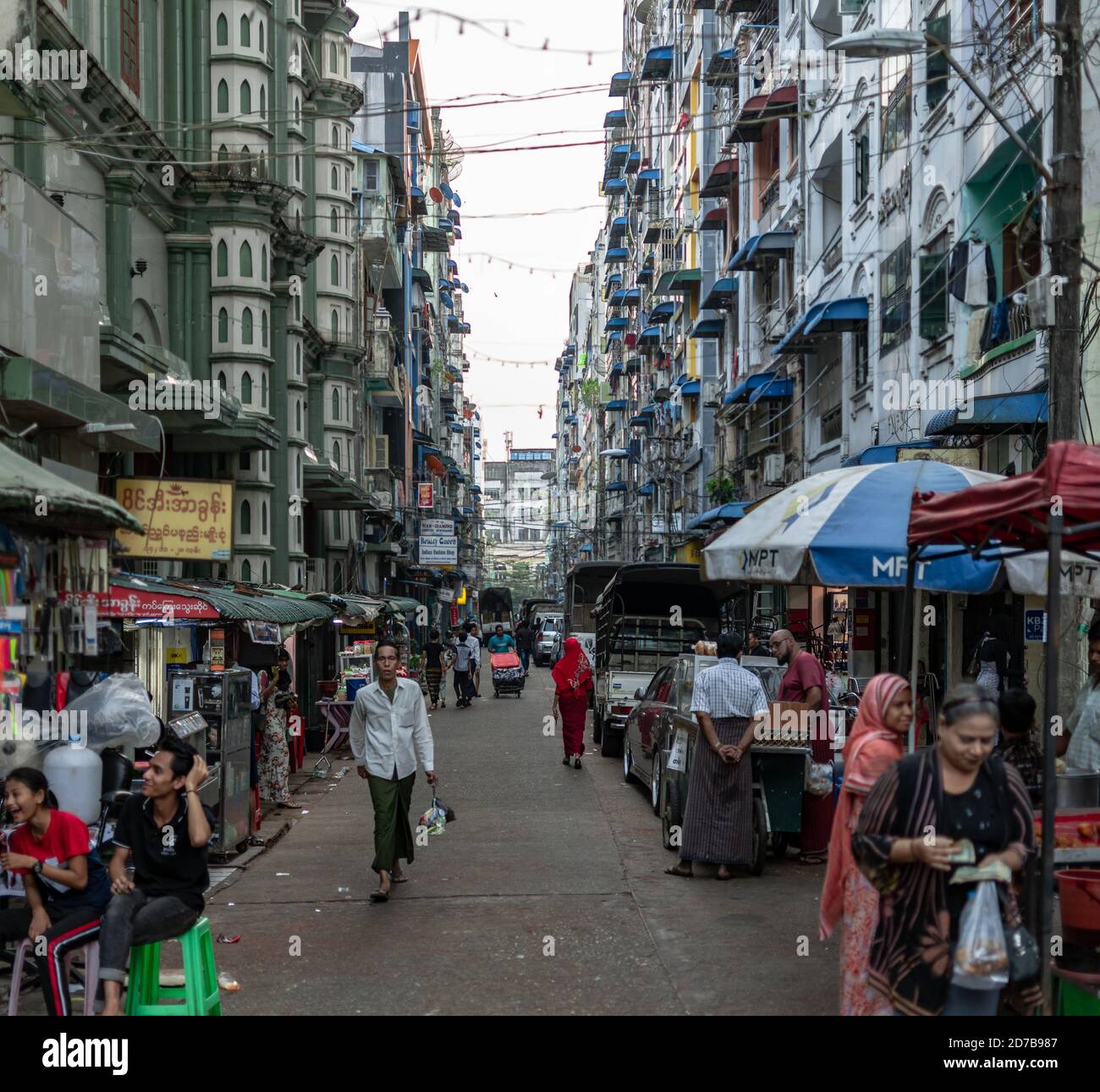 Yangon, Myanmar - 30 de diciembre de 2019: La vida cotidiana ocurre en una calle estrecha local en el centro de la ciudad con coches estacionados y peatones caminando Foto de stock