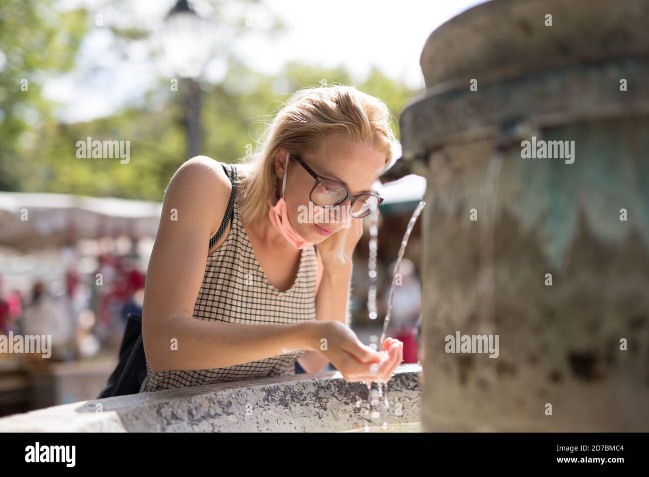 Mujer cucasiana joven y sedienta con mascarilla médica que bebe agua de la fuente de la ciudad pública en un caluroso día de verano. Nuevas normas sociales durante Foto de stock
