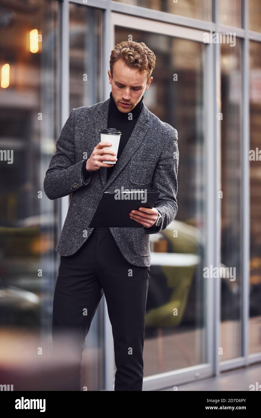 Hombre en elegante ropa formal con taza de bebida bloc notas en las manos está fuera contra el edificio moderno Fotografía de stock - Alamy