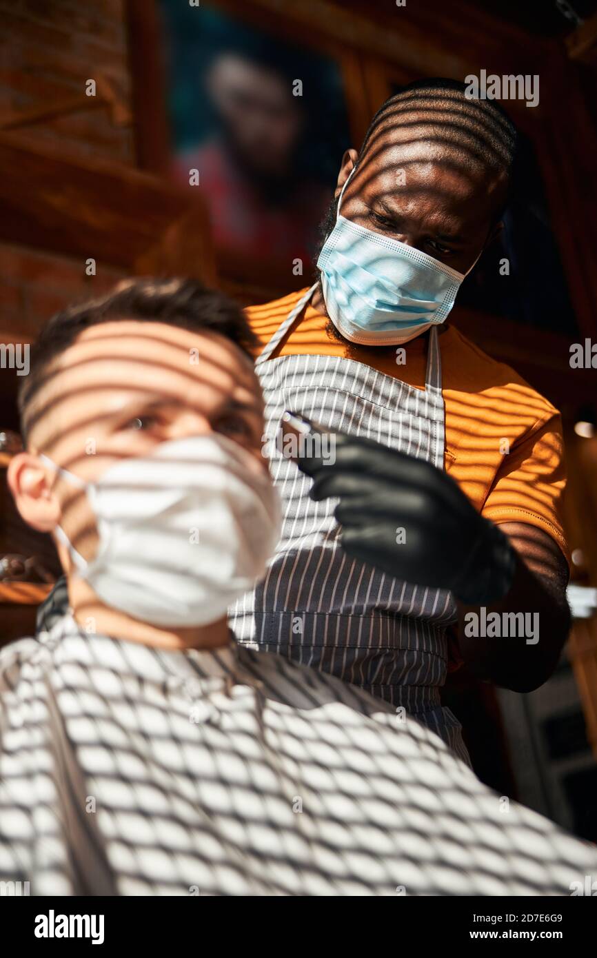 Barbería en máscara médica cortando el cabello de los clientes en la peluquería Foto de stock