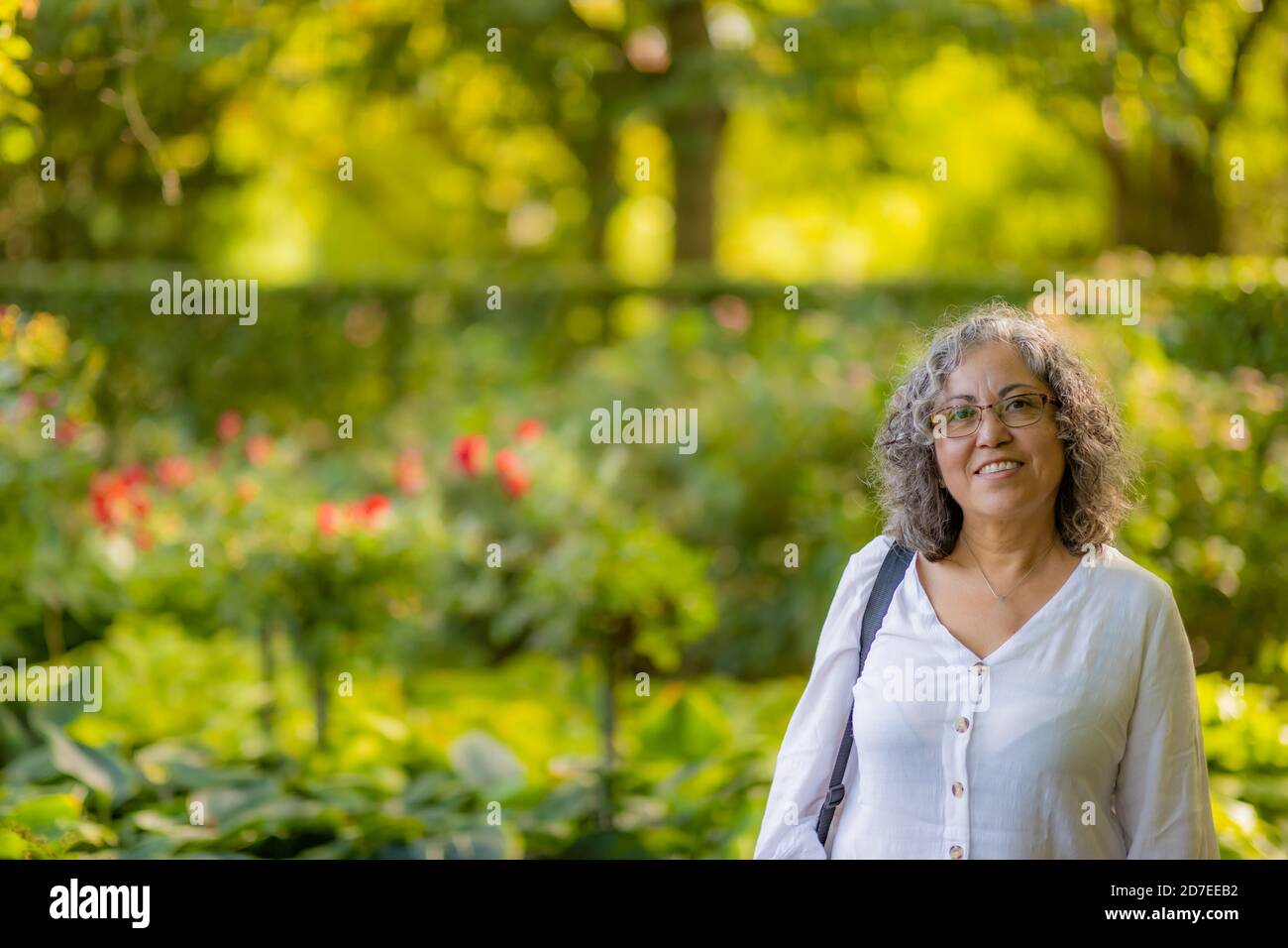 Sonriente mujer mexicana madura con gafas con ropa blanca casual con  exuberante vegetación verde y flores rojas en el fondo borroso, soleado  suma Fotografía de stock - Alamy