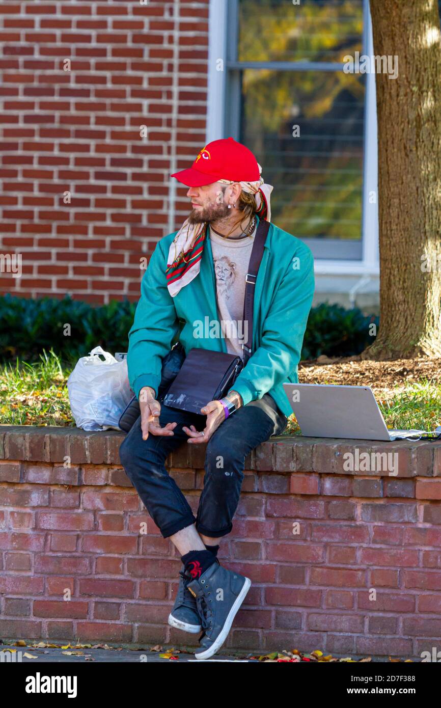 Frederick, MD, EE.UU. 10/14/2020: Un joven rubio libre de espíritu que lleva bandana bajo su sombrero de béisbol está sentado en una pared en la calle. Él tiene su l Foto de stock