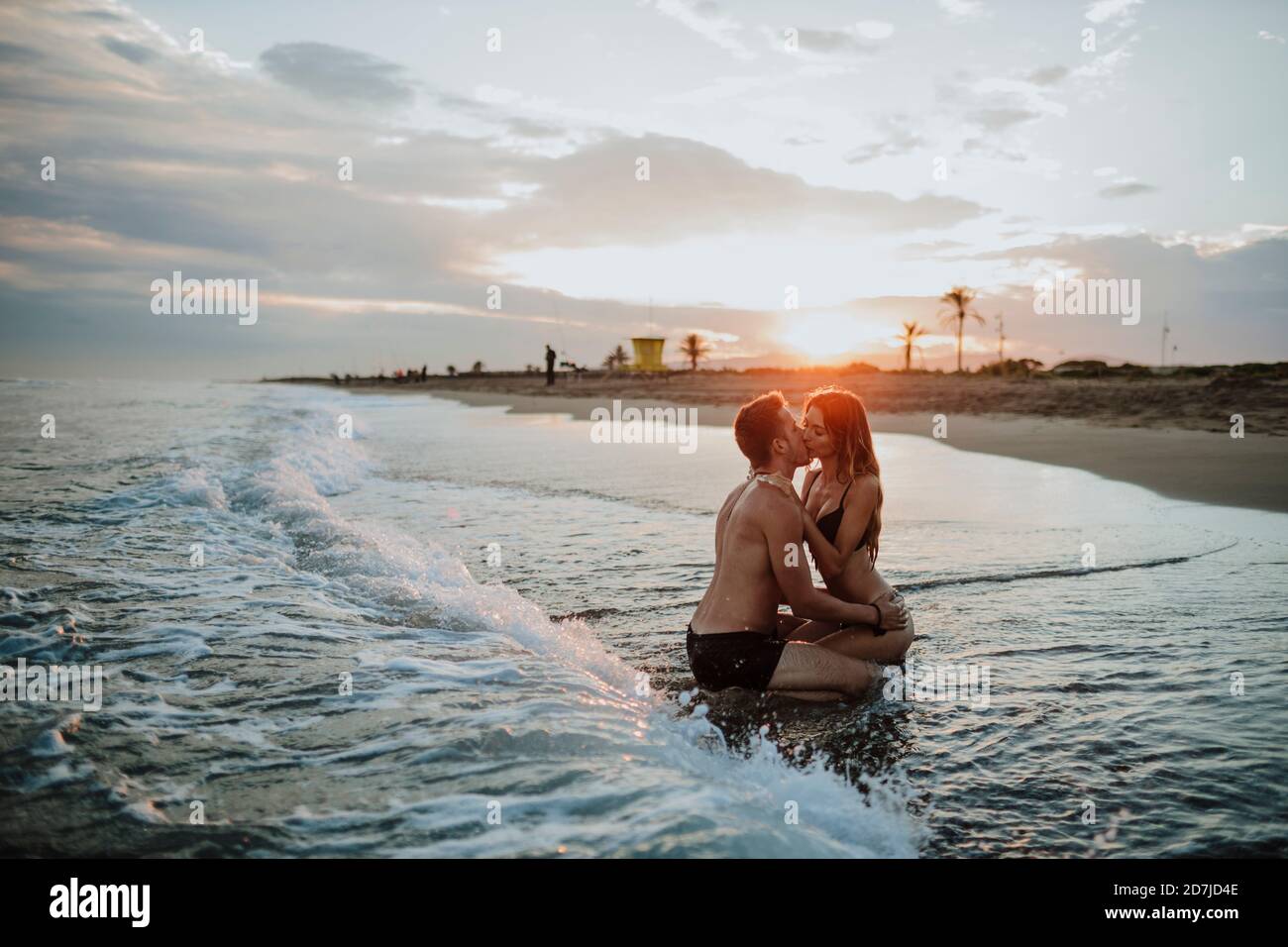 Pareja con traje de baño besándose mientras se sentaba en la playa durante  la puesta de sol Fotografía de stock - Alamy