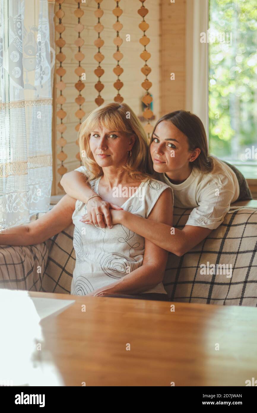 Amando a la hija de 20 años abrazar a la madre anciana desde atrás mientras mamá sentada en el sofá gente posando mirando la cámara sonriendo se siente feliz, concepto de mu Foto de stock