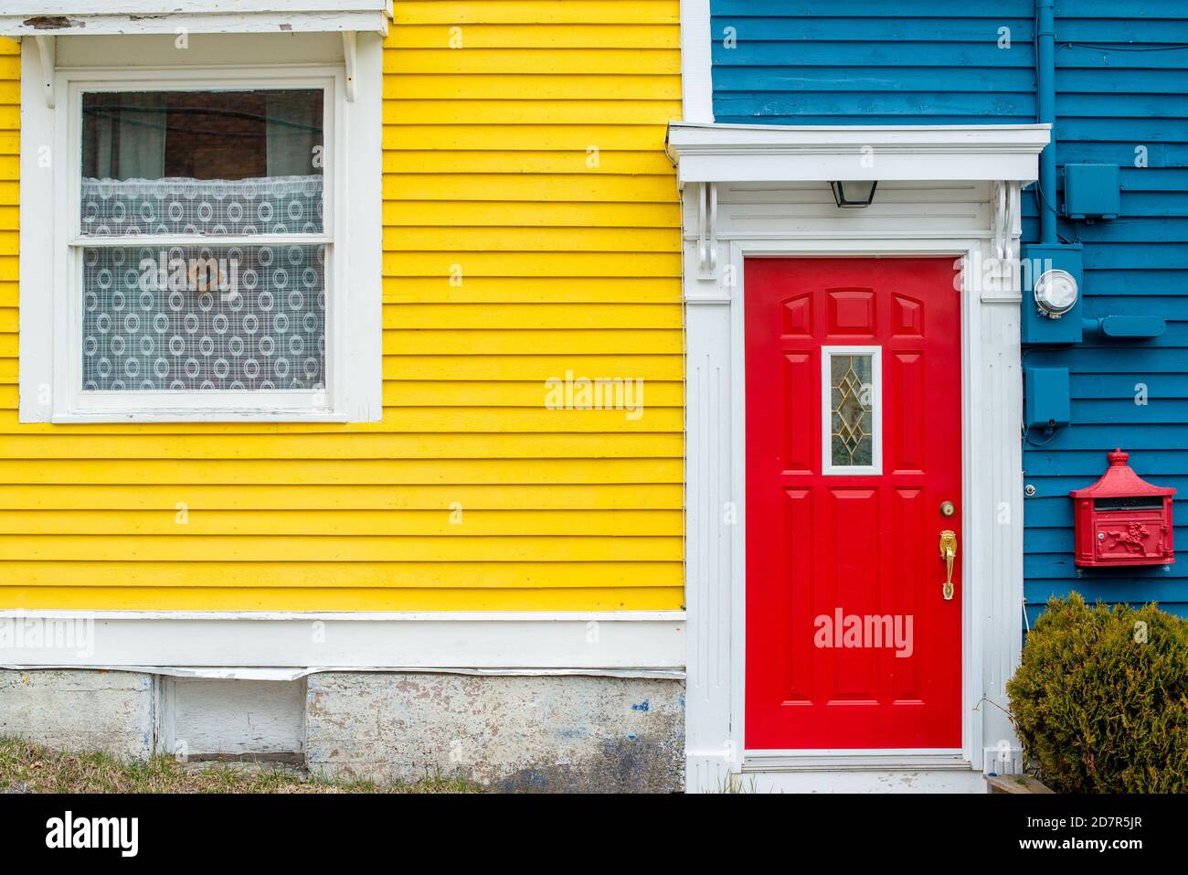 Una puerta roja brillante de un edificio con paredes de clapboard de madera azul y amarilla. La casa tiene un buzón de metal decorativo rojo. Foto de stock