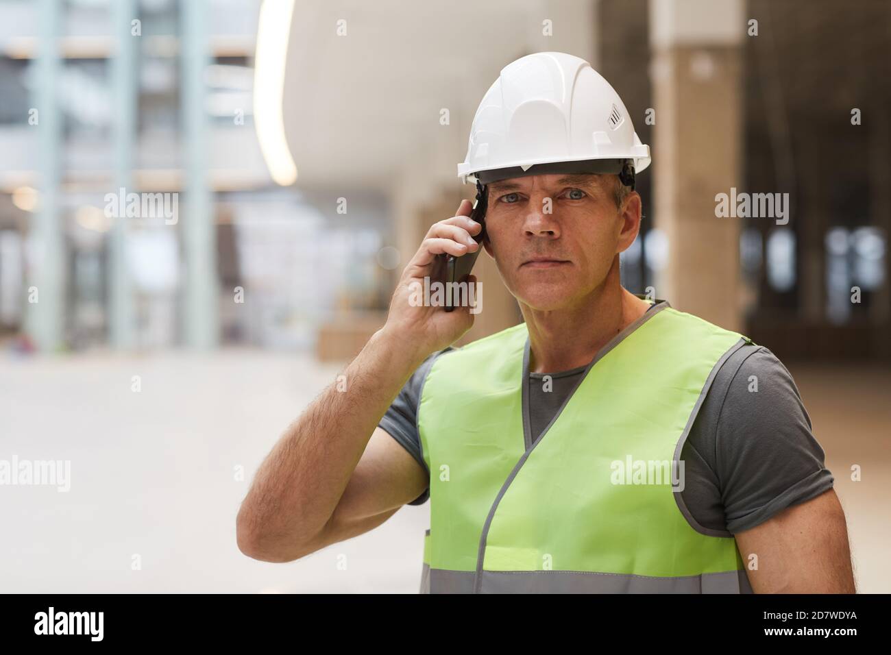 Retrato de un trabajador profesional de la construcción hablando por teléfono y mirando la cámara mientras está parado en el sitio de la construcción, espacio de copia Foto de stock