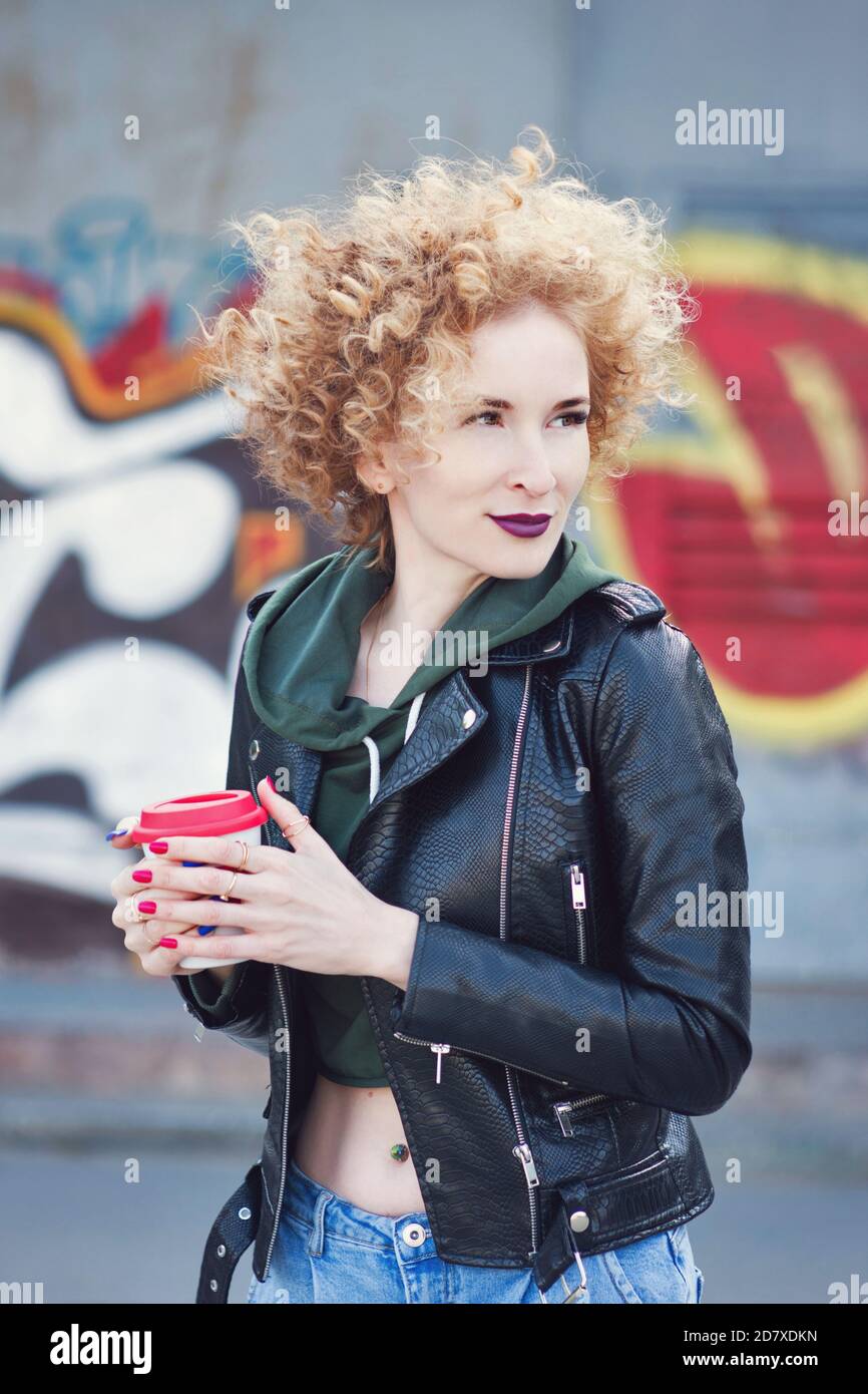 Cabello rojo rizado milenial chica que usa en estilo negro rock al aire libre incity cerca de la pared de graffiti. Hipster en la calle ropa de la celebración de taza de café. Juventud, estilo urbano, estilo de vida, concepto de tiempo libre. Foto de stock