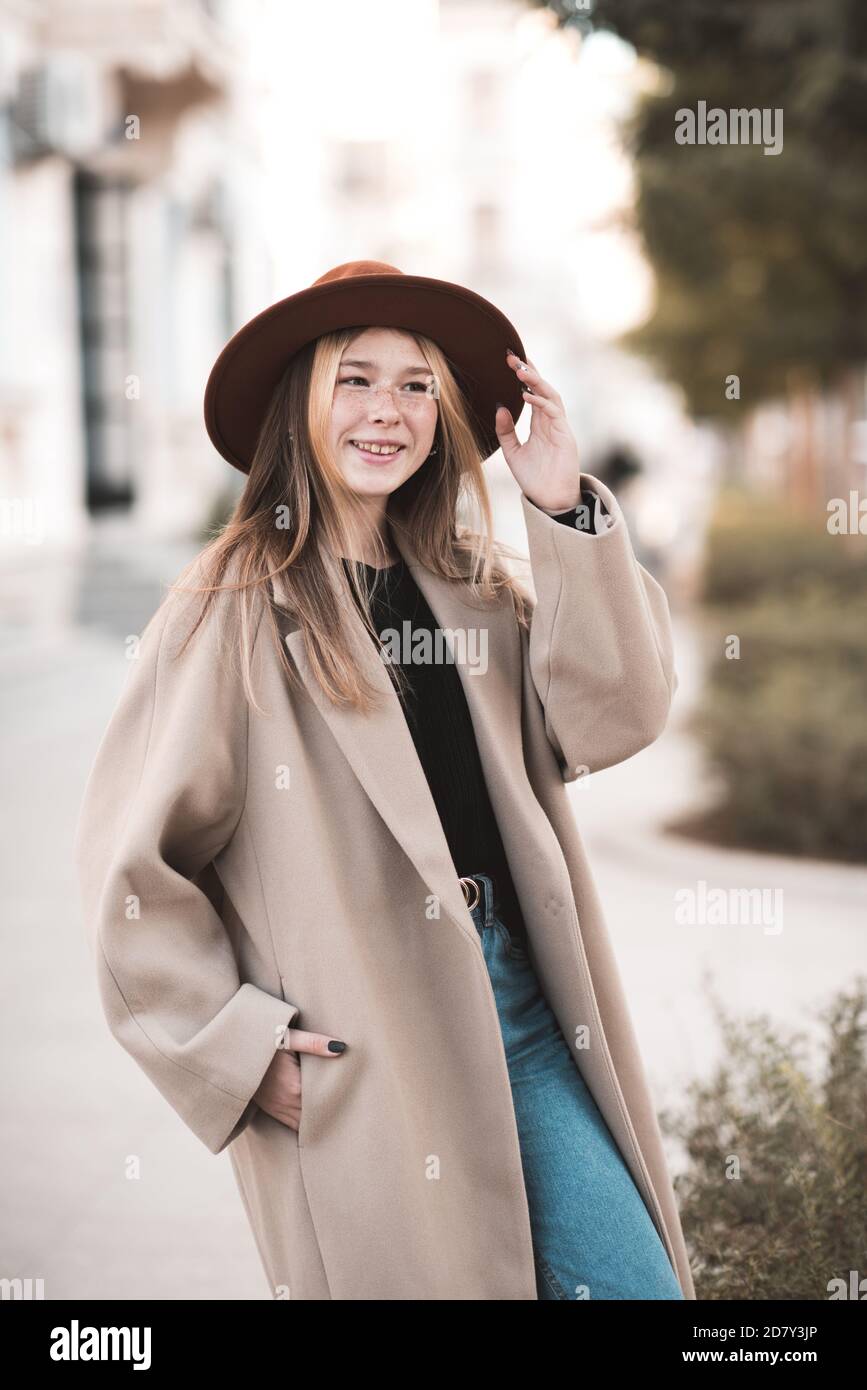 Chica adolescente sonriente de 14-15 años con sombrero y abrigo de invierno  caminando en la calle de la ciudad al aire libre de cerca. Mirando la  cámara. La capucha de la adolescente