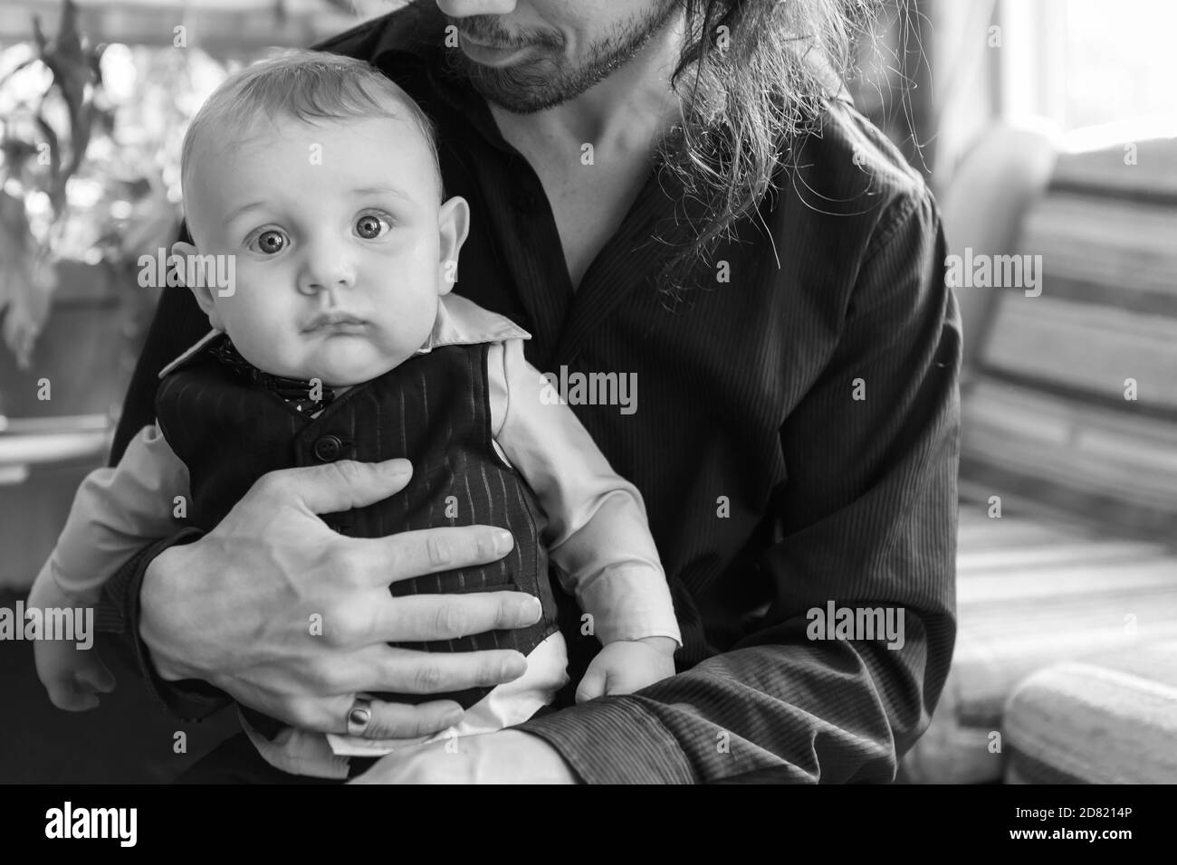 Retrato en blanco negro de un lindo bebé niño vestido, con ropa formal con lazo sentado en el hombre con un largo regazo de pelo en casa Fotografía de stock -