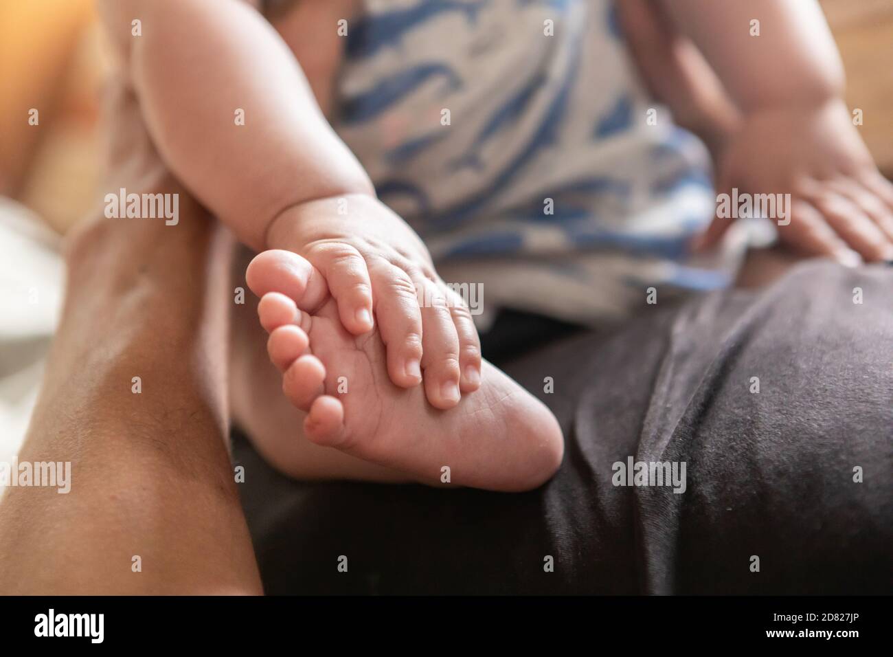 Enfoque selectivo de las manos que tocan las piernas y los pies del bebé niño mientras descansa y sentado en el vientre de su padre con apoyo en casa Foto de stock