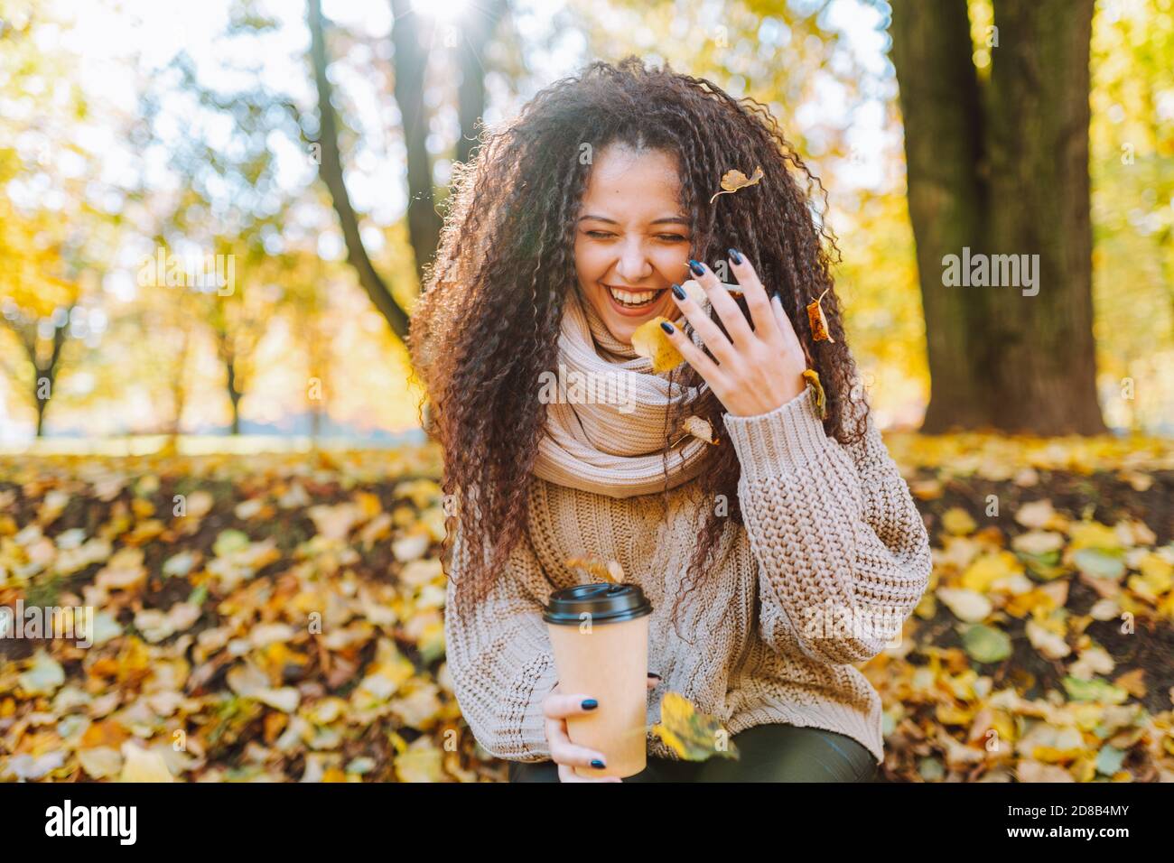 Positivo afro cabello mujer con hermosa sonrisa usando Jersey de punto y la bufanda tiran hojas amarillas de otoño en el parque al sol día Foto de stock