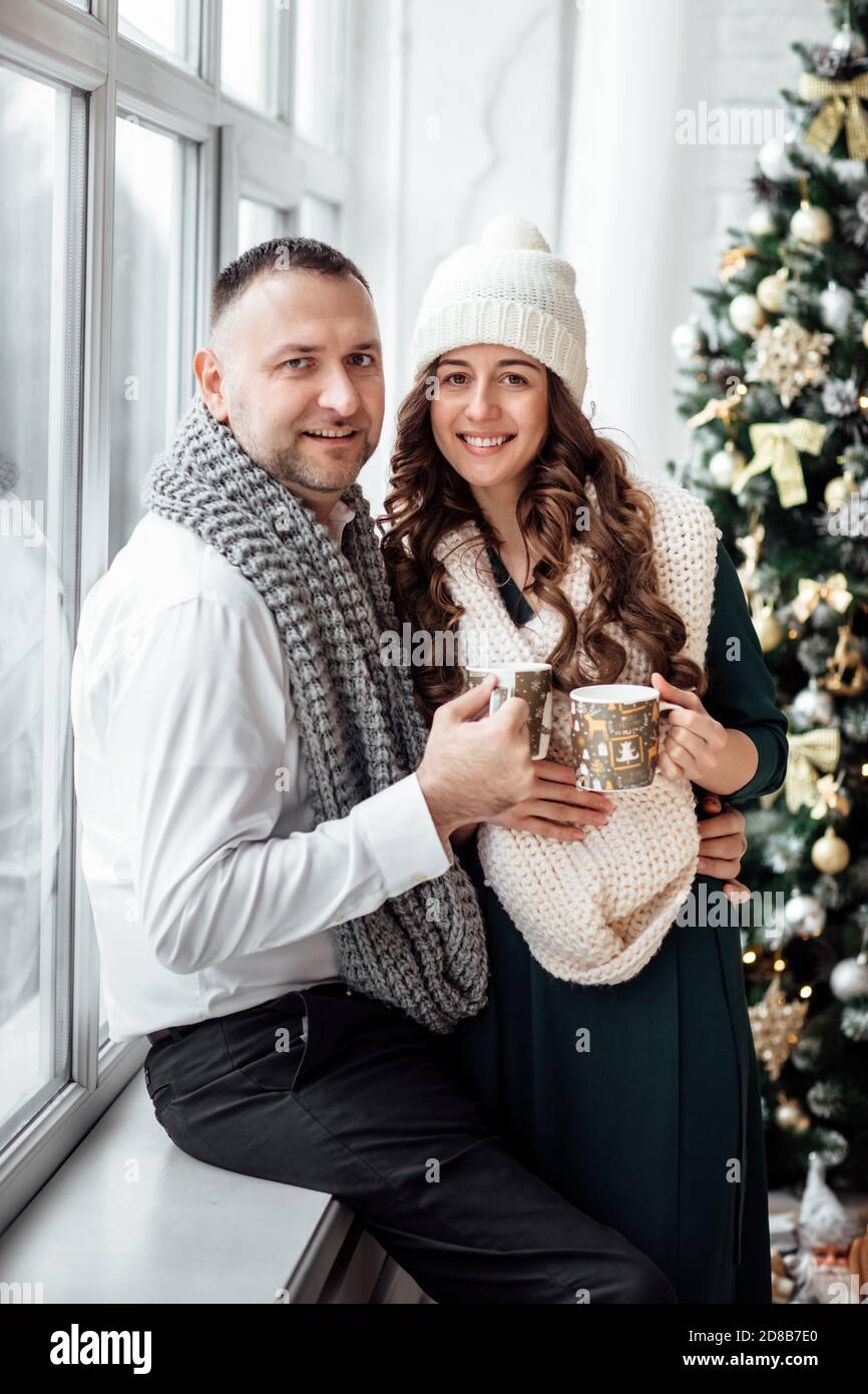 Navidad. Amor. Inicio. La pareja joven con ropa caliente está sosteniendo  tazas, hablando y riendo mientras se sienta en casa cerca del árbol de  Navidad Fotografía de stock - Alamy