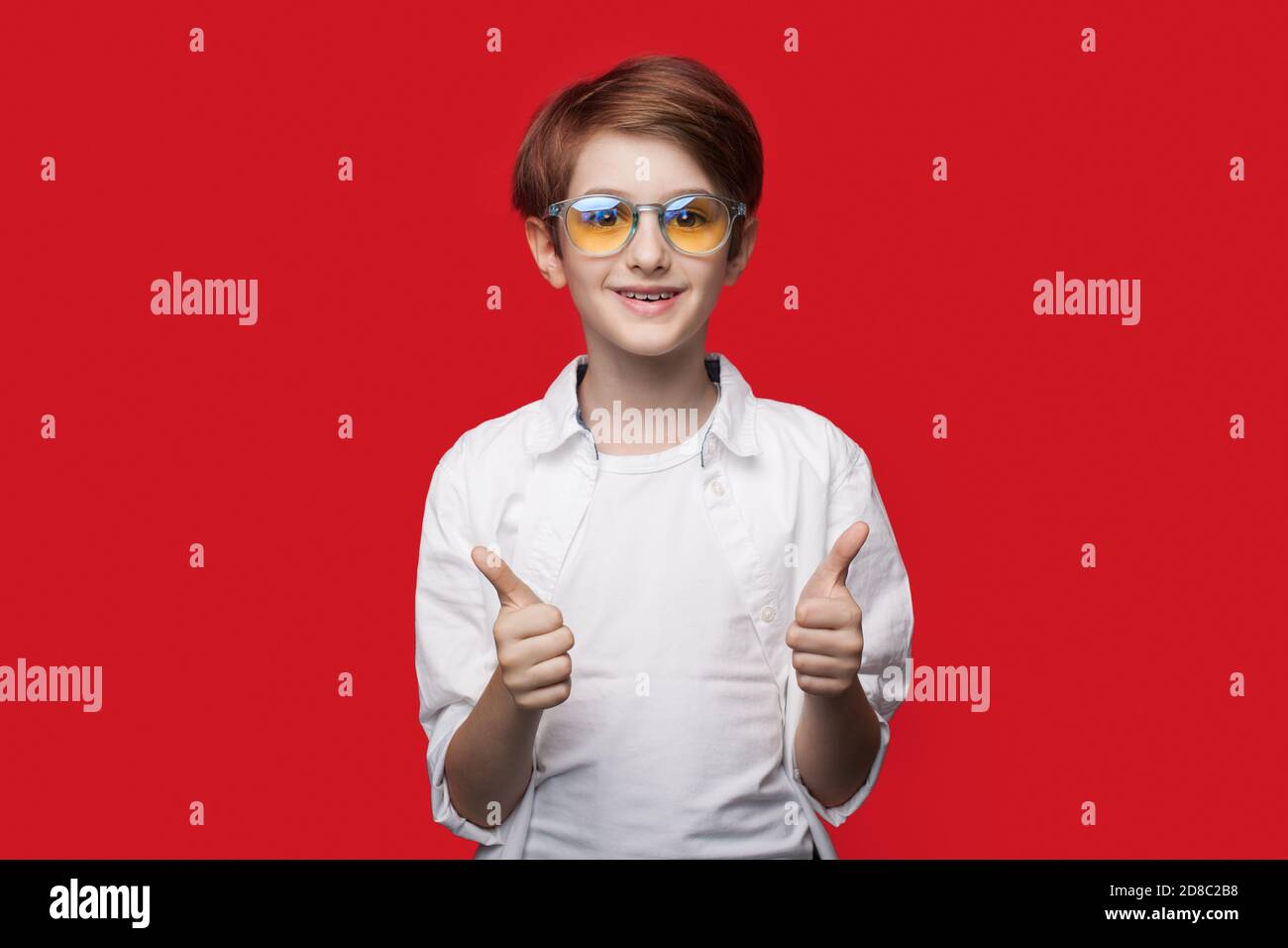 El joven caucásico con gafas está gestando el signo similar en una pared roja del estudio sonriendo a la cámara Foto de stock