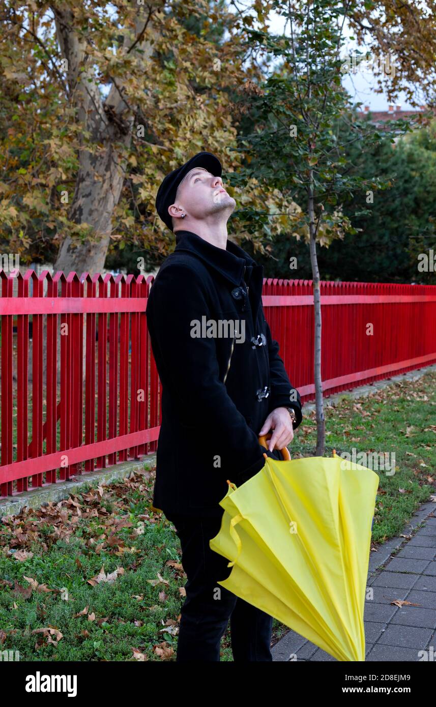 Hombre en abrigo negro, de pie en las calles de la ciudad con paraguas  amarillo en sus manos, mirando al cielo para las gotas de lluvia.  Pronóstico y tiempo lluvioso en otoño