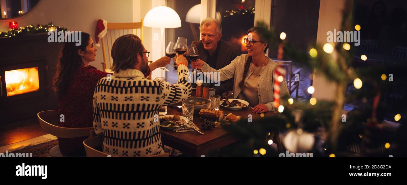 Familia europea sentada en la mesa de la cena tostar vino en la víspera de Navidad. La familia tiene una cena de acción de gracias en casa. Foto de stock