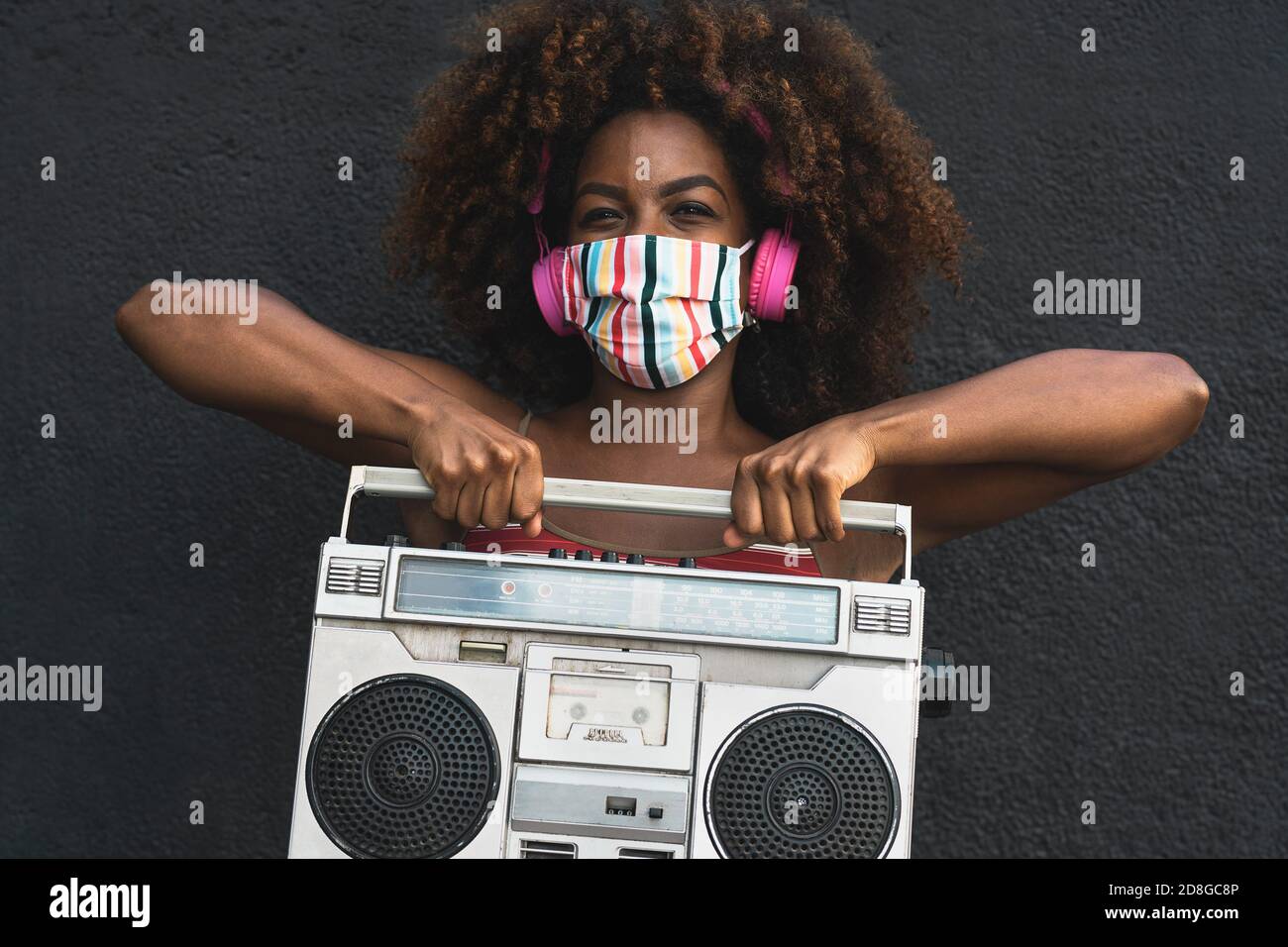 Joven mujer afro con máscara bailando al aire libre mientras escucha a la música con auriculares inalámbricos y boombox clásico estéreo Foto de stock