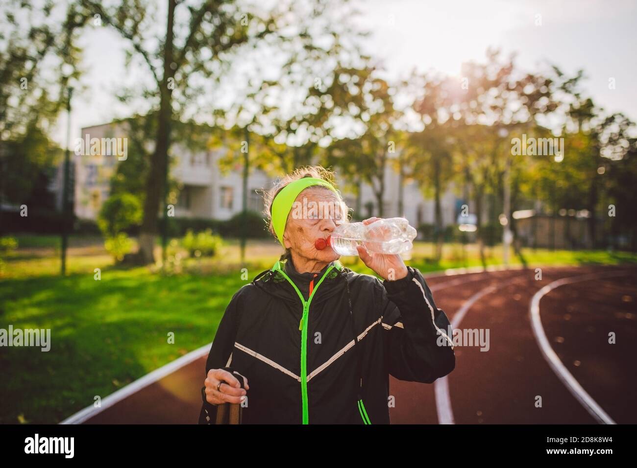 Mujer mayor practicando marcha nórdica en el estadio de la ciudad y se detuvo para saciar la sed, beber agua de la botella. Edad, madurez, estilo de vida activo y. Foto de stock
