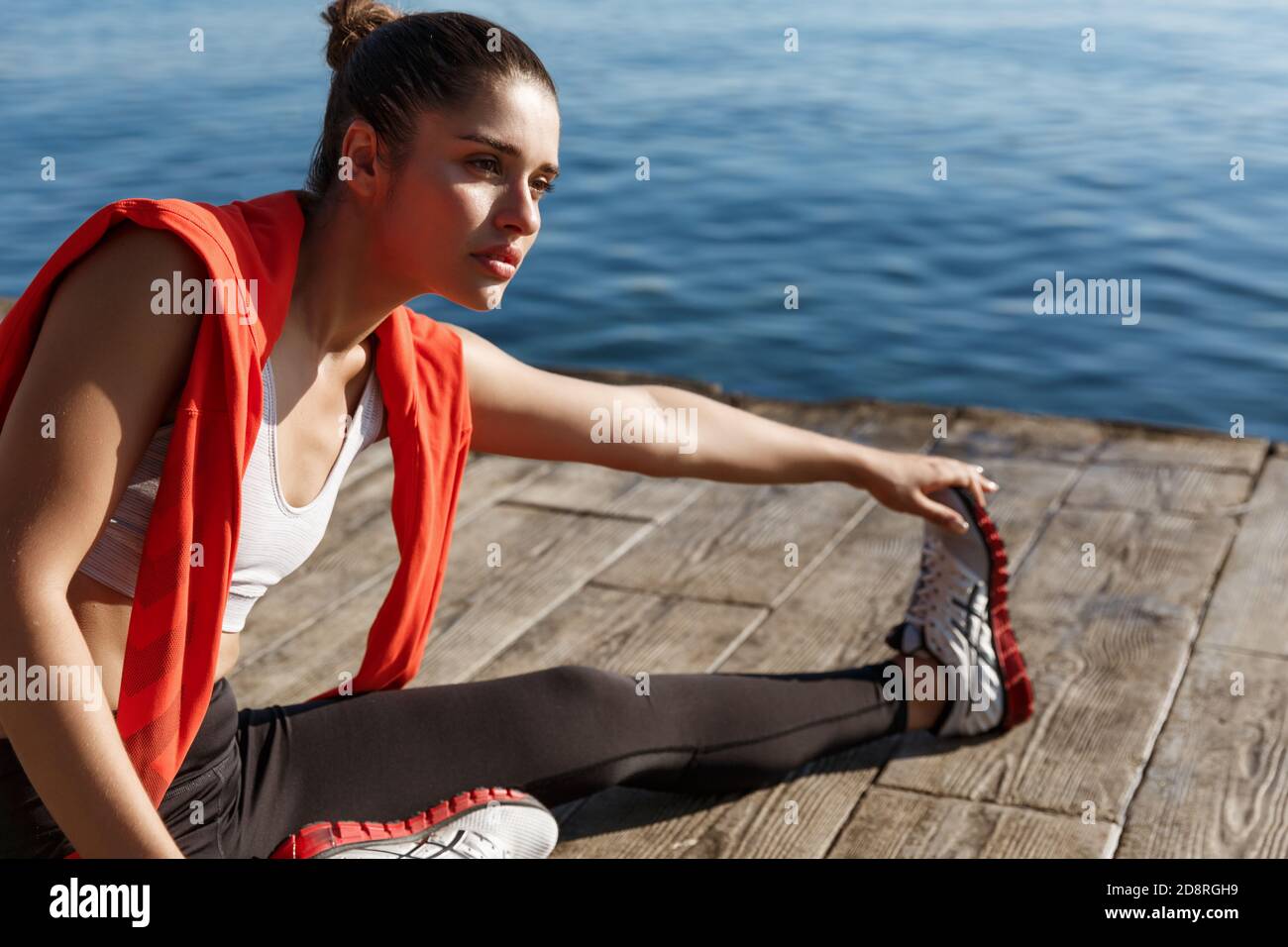 Foto al aire libre de atractiva mujer de fitness calentándose antes de correr, sentado en el muelle y estirando las piernas Foto de stock