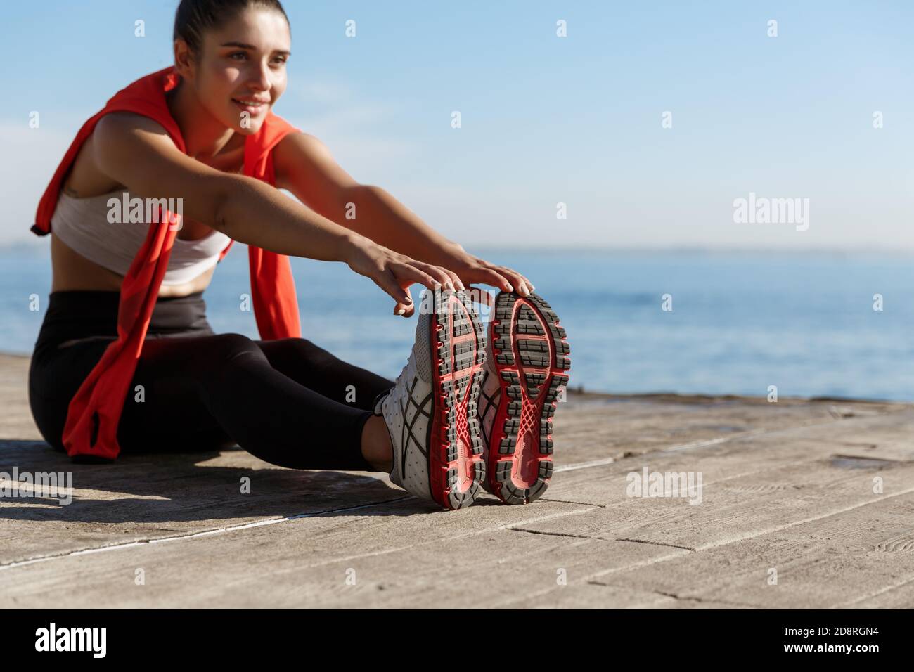 Tiro al aire libre de sonriente entrenamiento deportivo seguro cerca del mar. Mujer de fitness sentada en el muelle de madera y estirando las piernas Foto de stock
