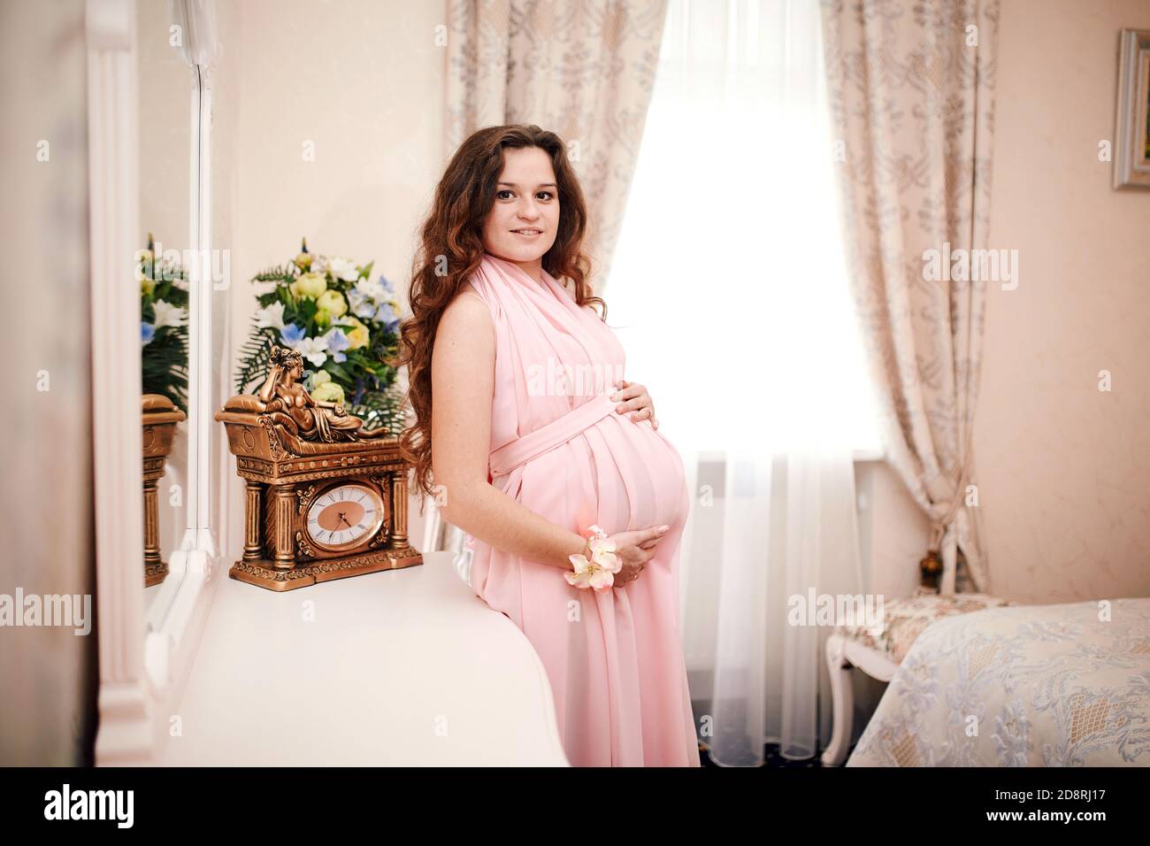 mujer de pelo rojo con un vestido rosa posando en un interior clásico a la esperando a un bebé. embarazo Fotografía de stock -