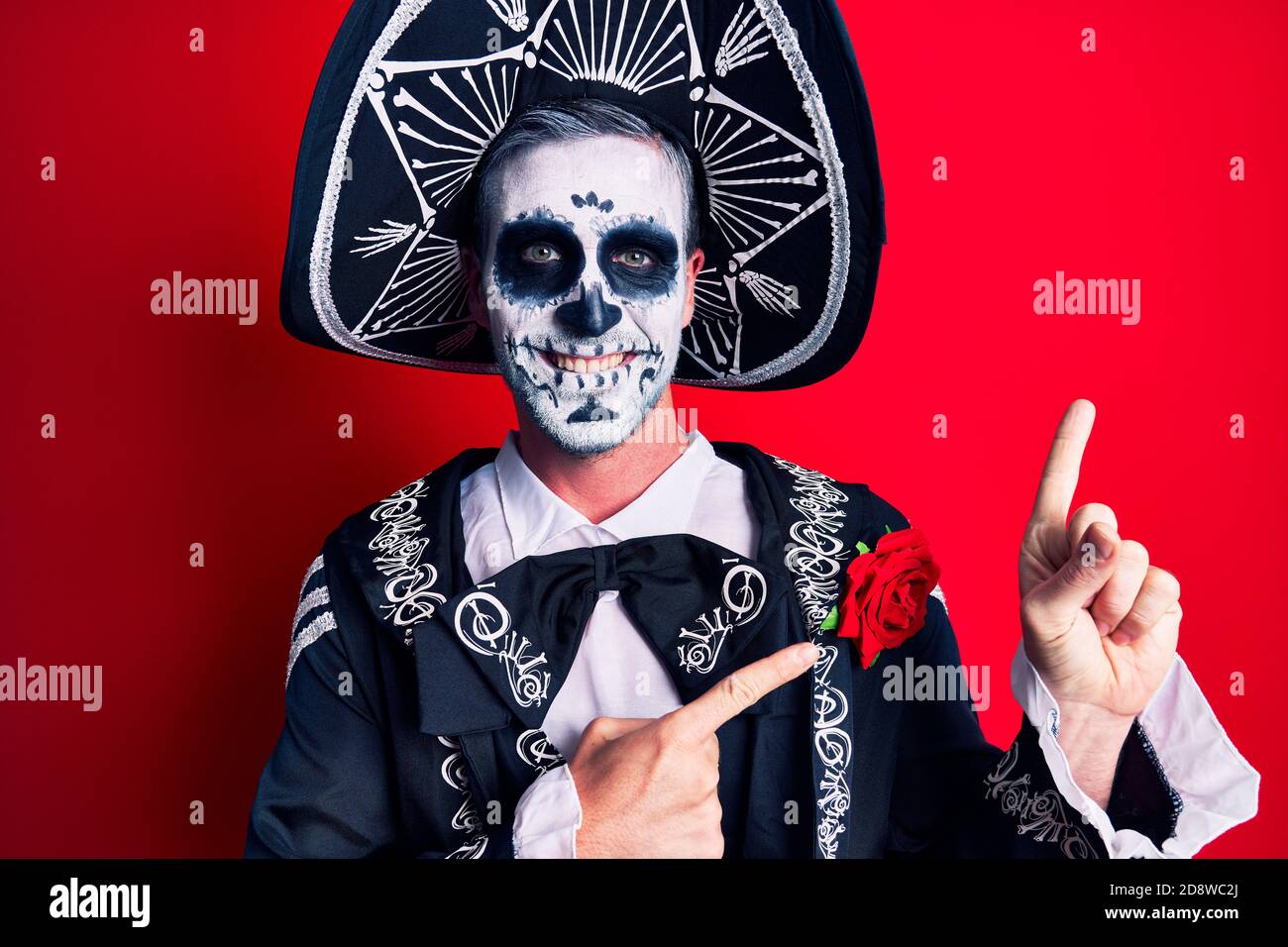 Un joven que viste el día mexicano del traje muerto con una sonrisa roja y mirando la cámara apuntando con dos manos y dedos a un lado. Foto de stock